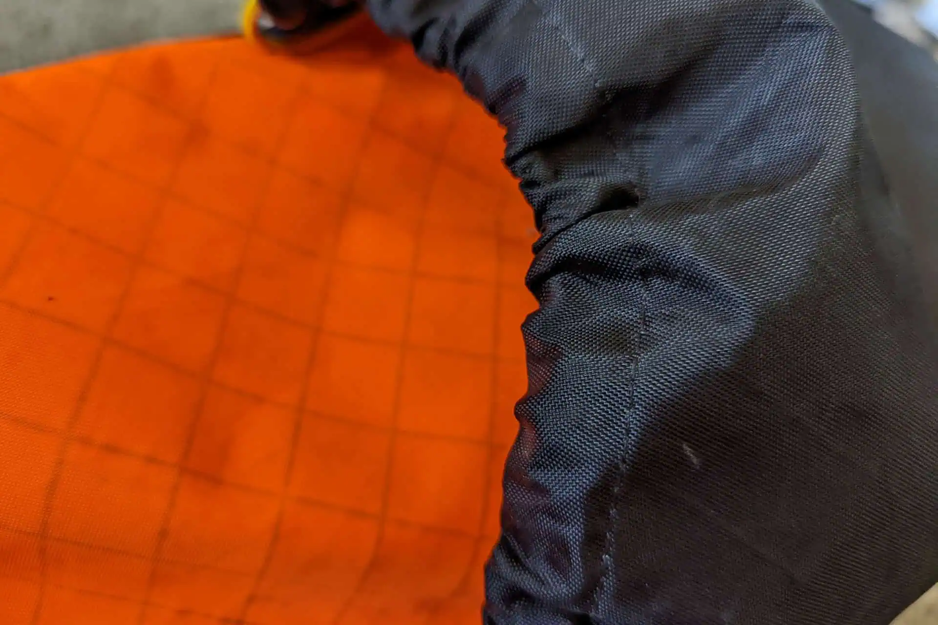 Close-up of an Atom Packs Pulse orange fabric with grid lines, partially covered by a black elastic material on the right. The textured fabric suggests it might be part of a bag or clothing item.