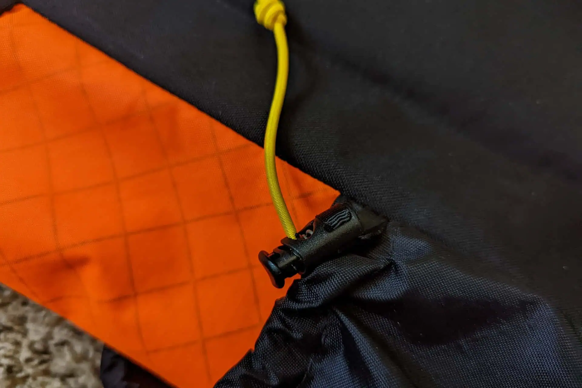 A close-up of a black drawstring toggle on a bright orange Atom Packs Pulse fabric with a grid pattern. A yellow cord threads through the toggle, highlighting part of the black and orange textile.