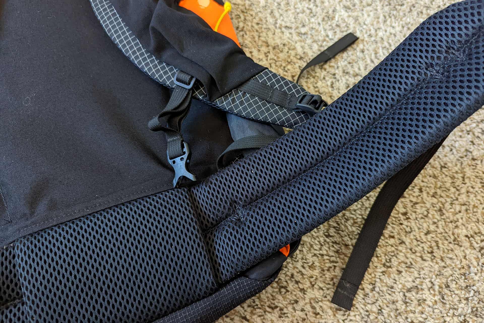Close-up of an Atom Packs Pulse black backpack on a carpeted floor. The image highlights the padded shoulder strap, showcasing its mesh fabric and adjustment buckle. Part of the backpacks main body is also visible.