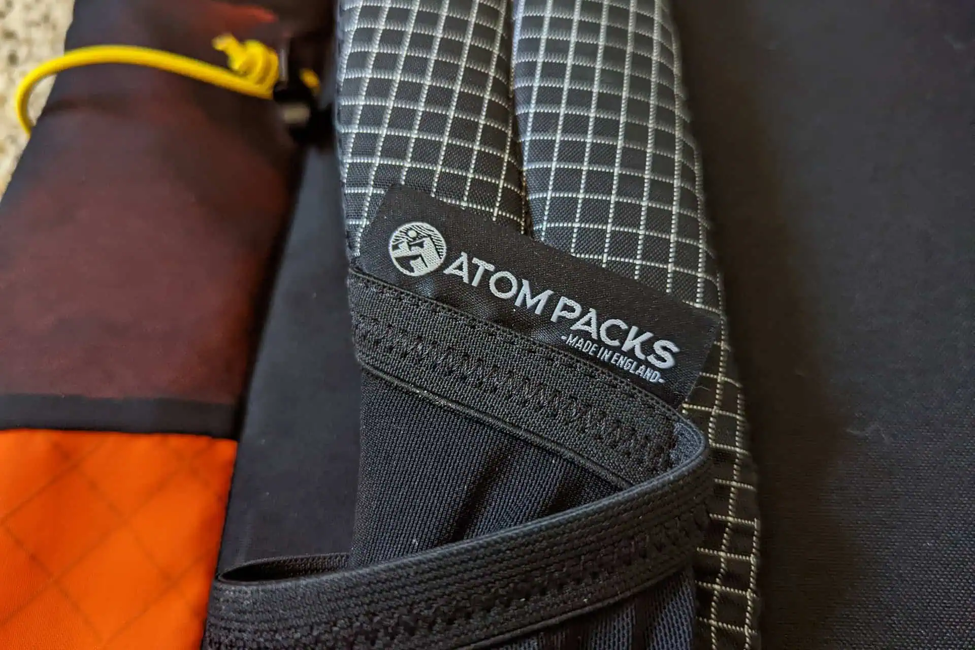 Close-up of a backpack showcasing the Atom Packs Pulse label with Made in England text. The material displays a checkered pattern and black color, accented by vibrant red and yellow tones.