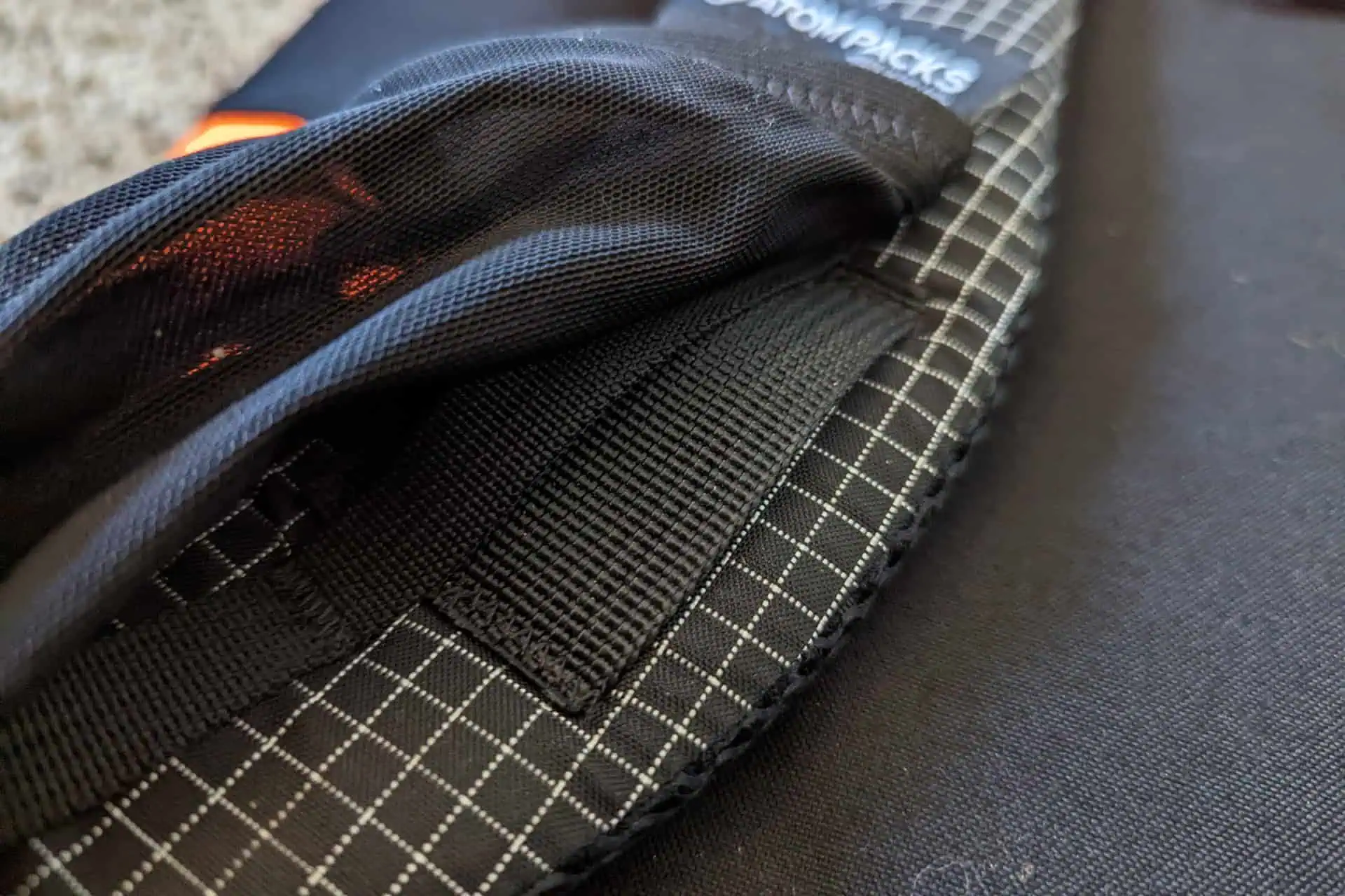 Close-up of a black Atom Packs Pulse backpack featuring a grid pattern and mesh fabric. A sturdy black strap is visible, with a hint of an orange object in the background.