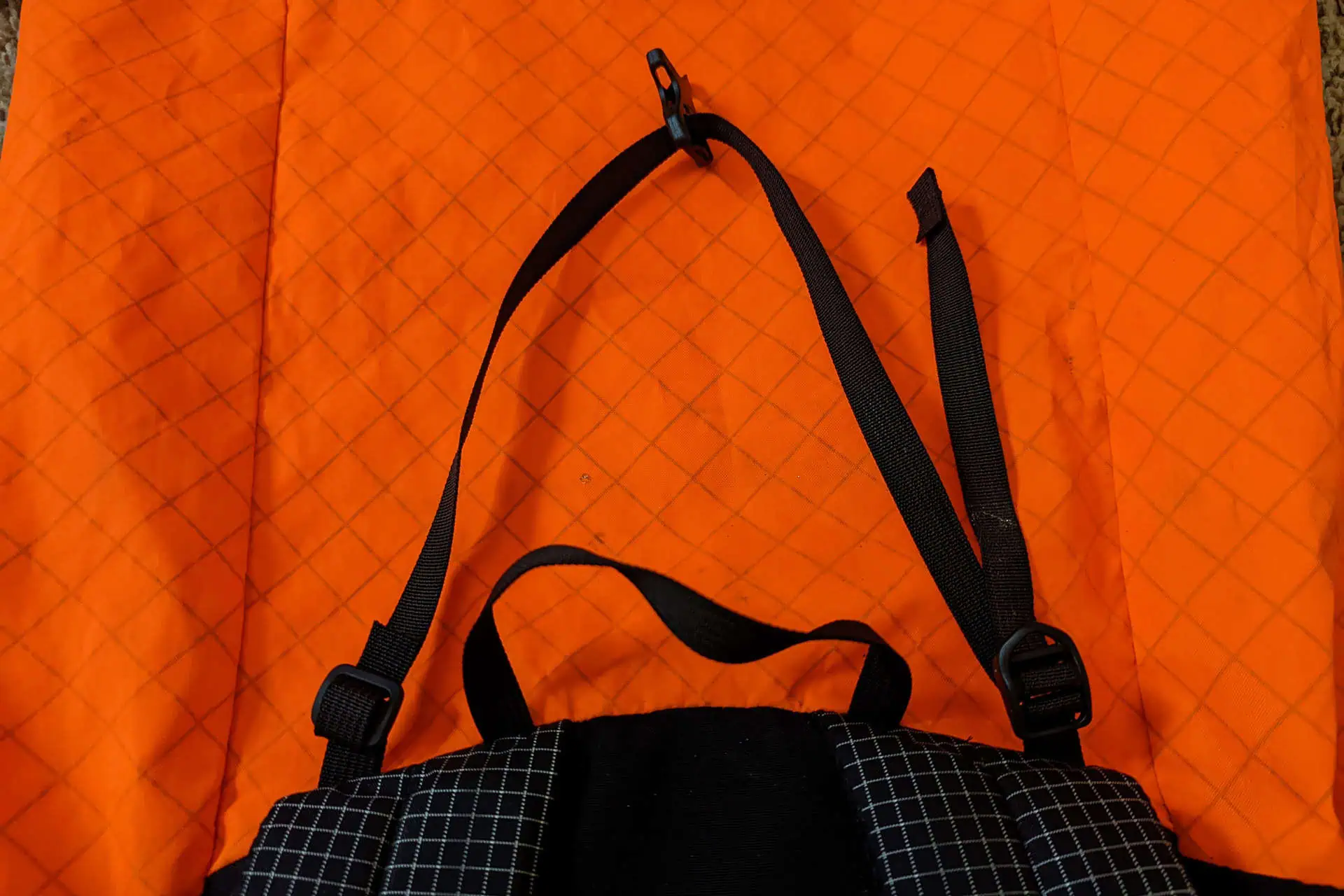 A black backpack strap from Atom Packs Pulse, featuring adjustable buckles, is laid on an orange quilted fabric with a diamond pattern. The straps clip at the end stands out against the bright, contrasting background.