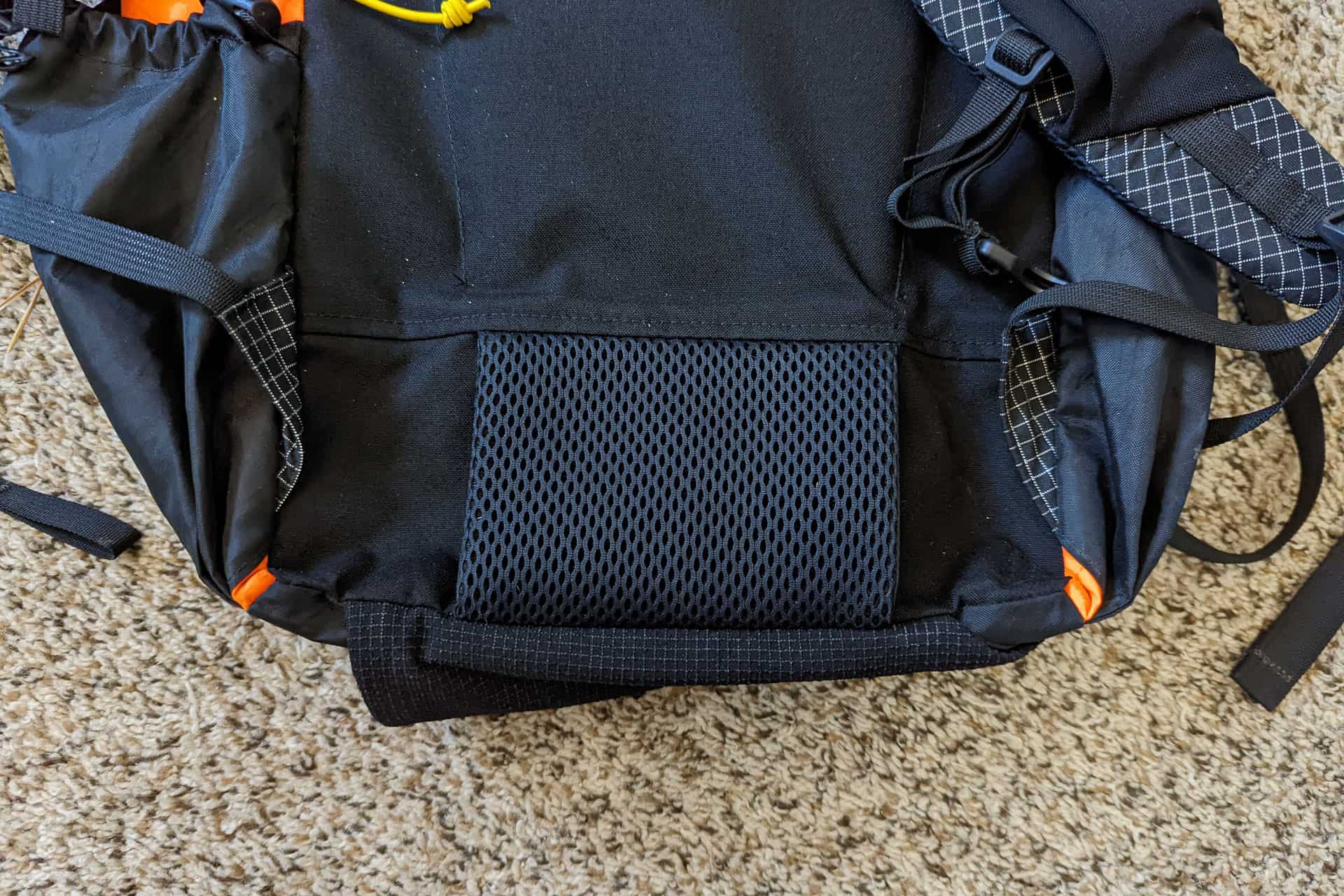 A close-up of an Atom Packs Pulse backpack resting on a carpeted floor. The black backpack features orange details, black mesh pockets, and adjustable straps. Textured fabric panels are visible along with some loose straps.