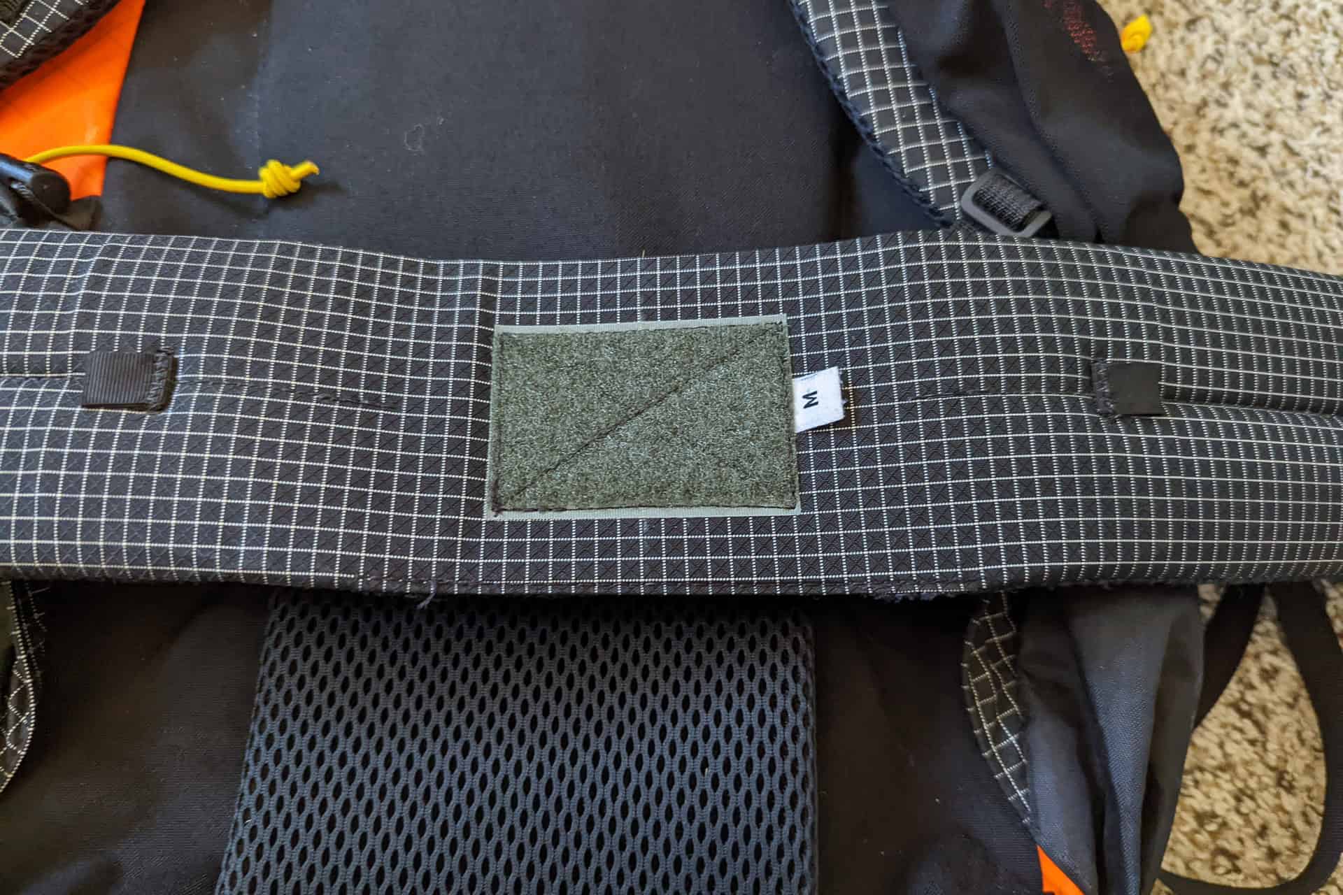Close-up of an Atom Packs Pulse backpack showcasing a grid-patterned strap with a size tag marked M. The black and white stitching features a Velcro panel, while the backpack’s black fabric is accented by hints of orange and a distinctive yellow cord.