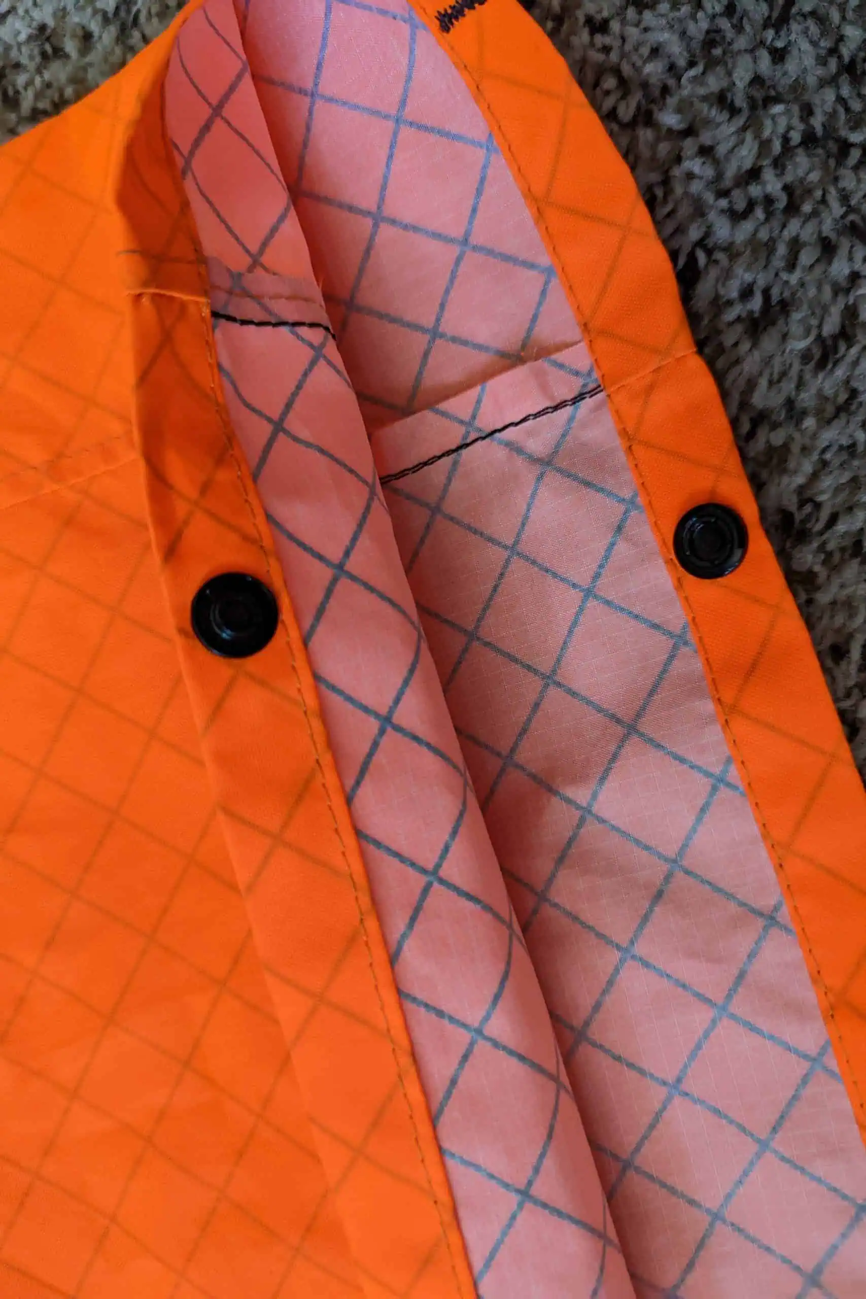 Close-up of an orange Atom Packs jacket with black buttons. The interior features a light pink lining with a blue diamond pattern, akin to the dynamic Pulse collection. The jacket is lying on a textured gray surface, showcasing its vibrant details.