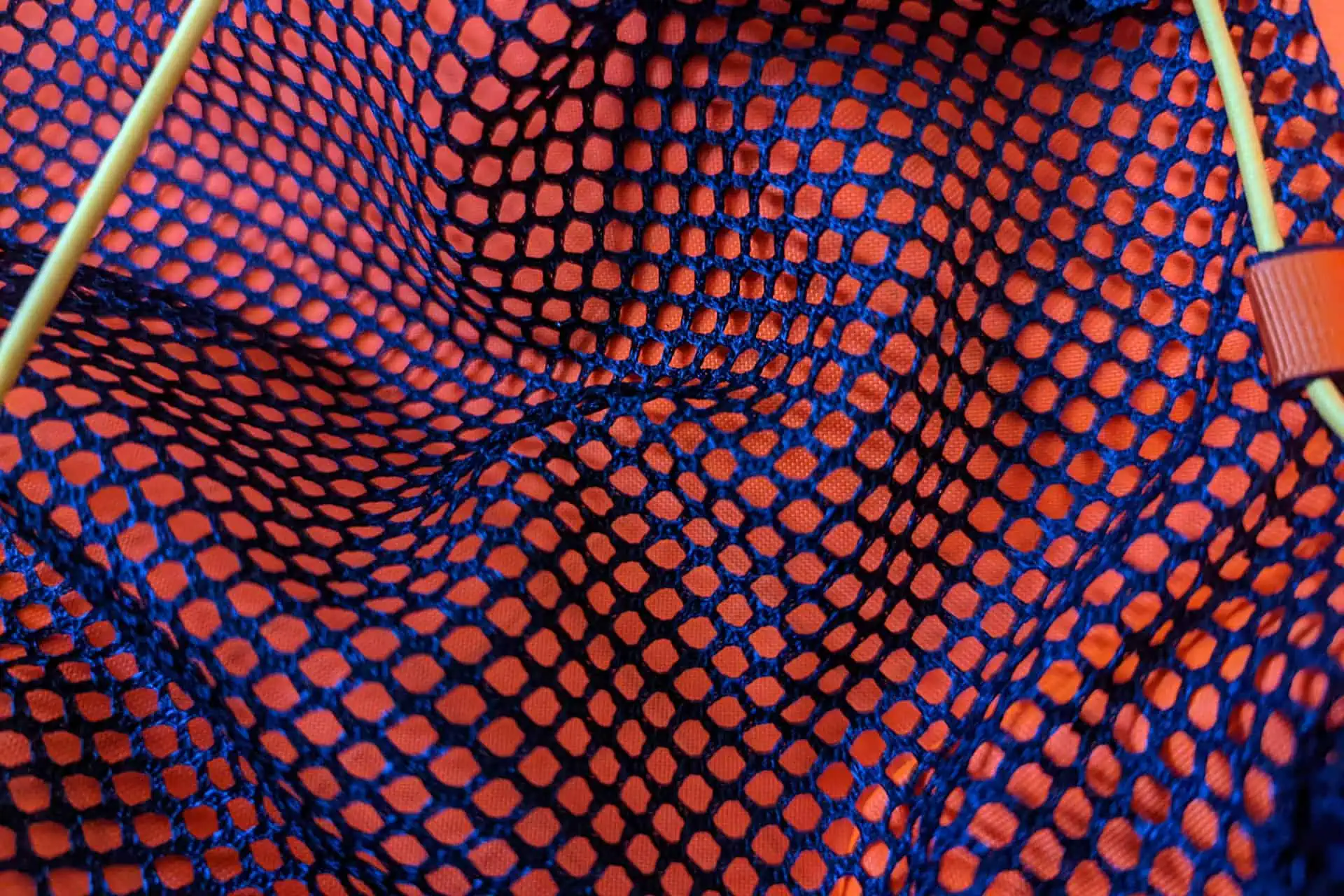 Close-up of an orange fabric with a blue net-like pattern creating a geometric, tactile texture reminiscent of Atom Packs. The fabric is slightly wrinkled, and light green rods at the corners hint at structure or support, pulsating with energy.