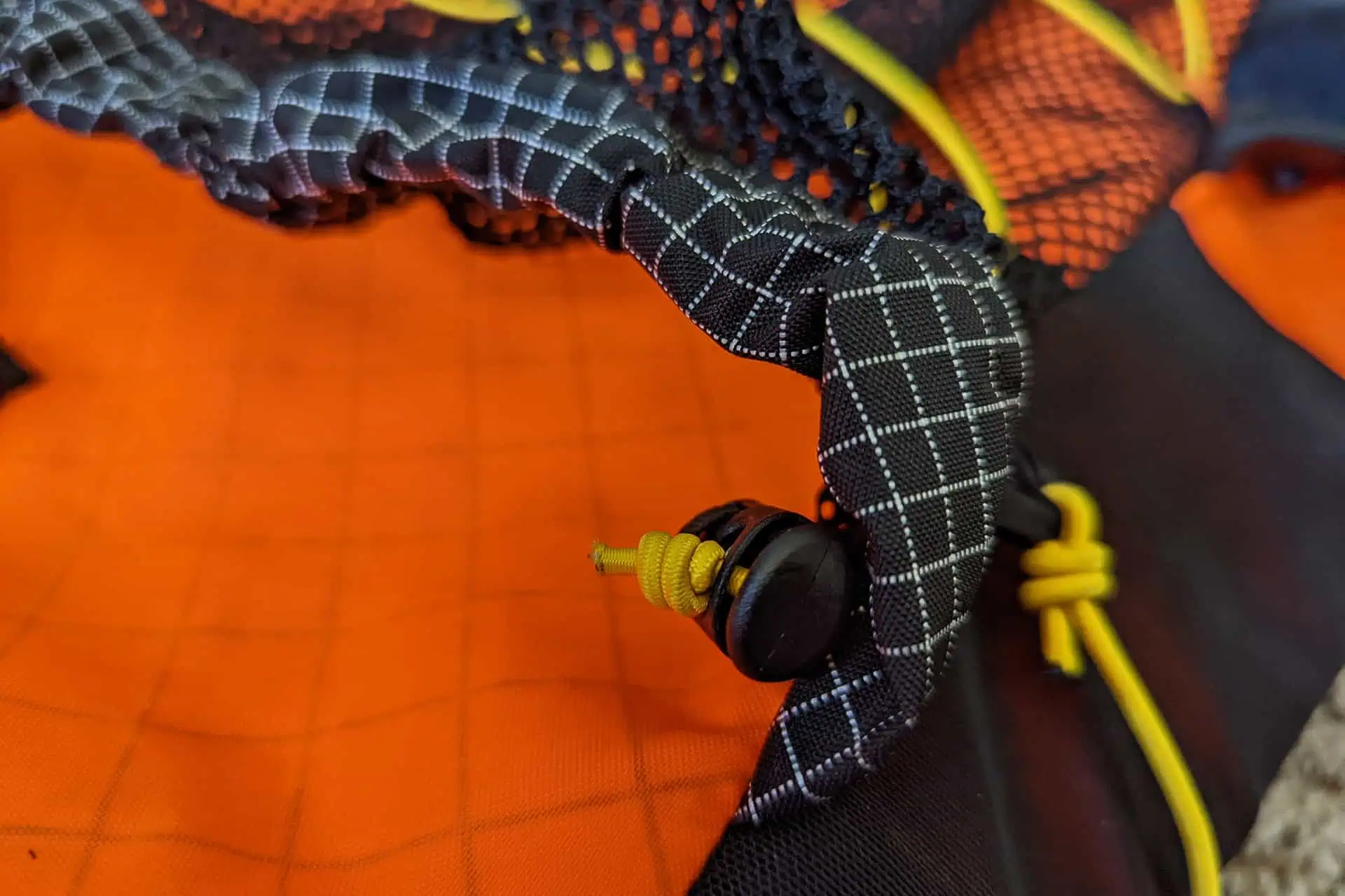 Close-up of an Atom Packs Pulse gear item, featuring an orange mesh fabric with black and white grid-patterned straps. A small yellow cord with a black toggle is visible.