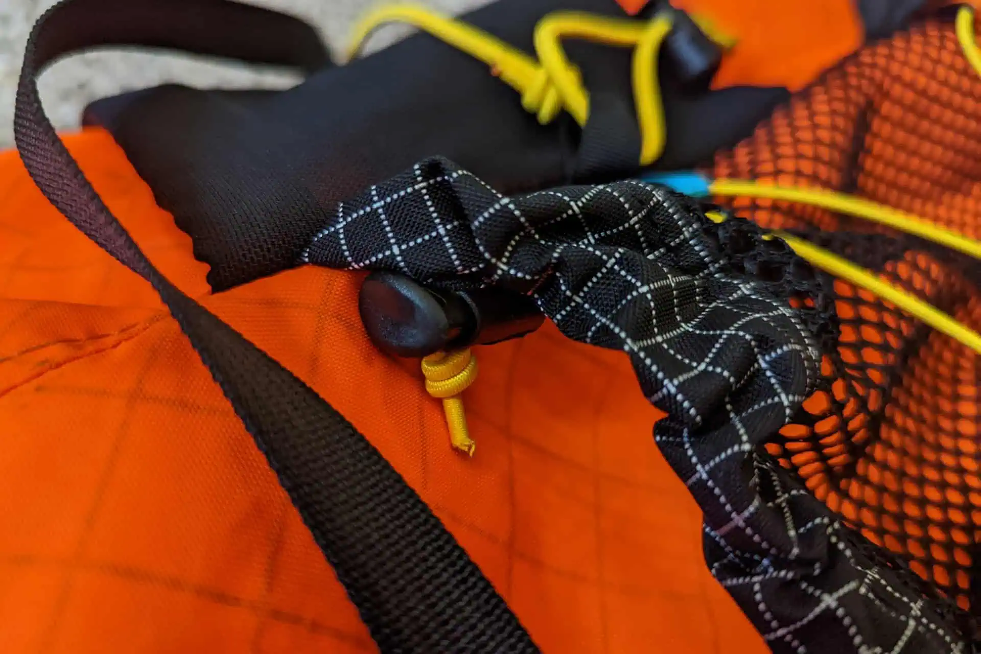 Close-up of an Atom Packs Pulse in vibrant orange with sleek black straps. A yellow-corded drawstring, cinched by a sturdy black toggle, stands out against the white grid pattern. A mesh section accentuated by yellow cords completes this eye-catching design.