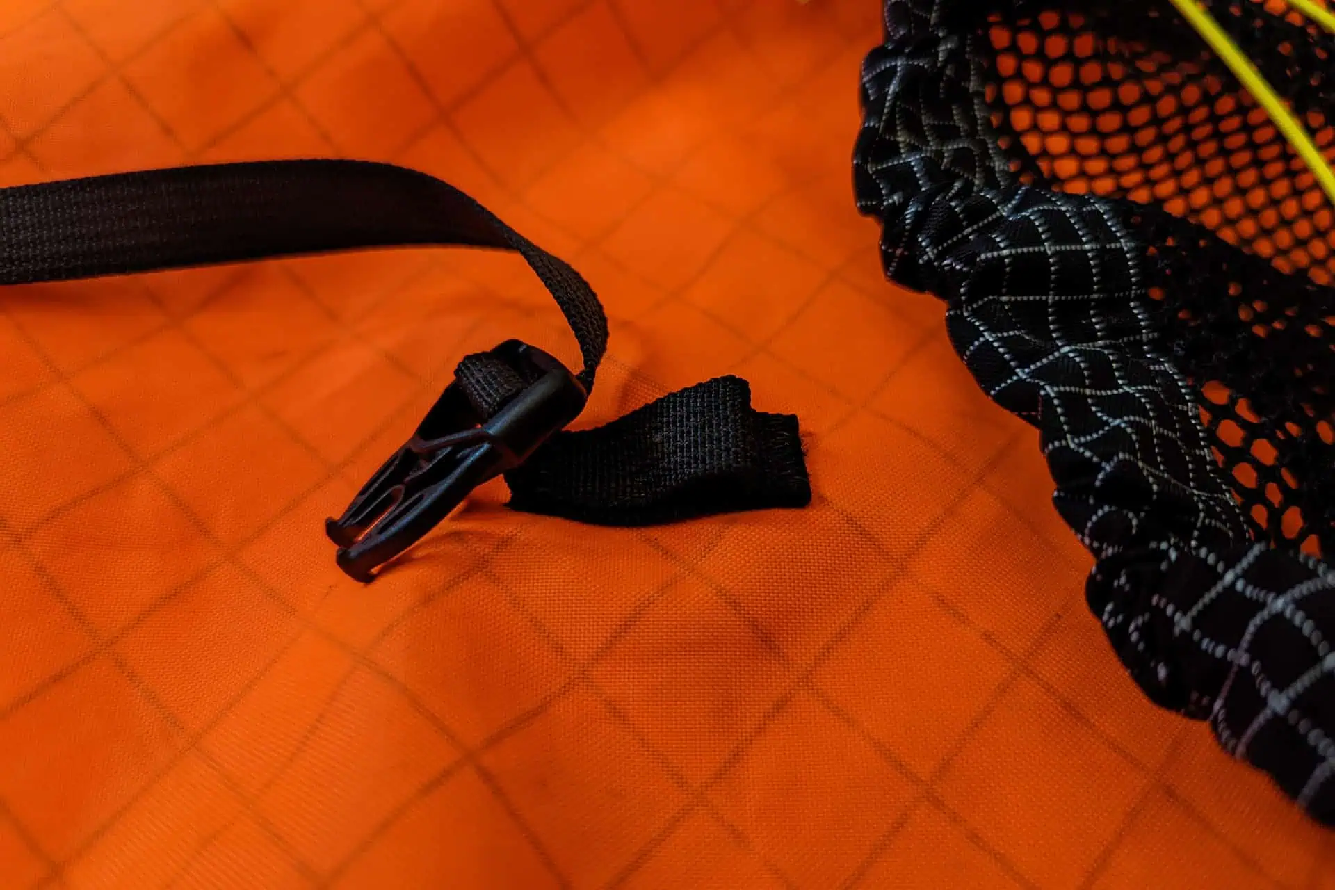 Close-up of Atom Packs Pulse design showcasing an orange fabric with a grid pattern, complemented by a black strap with a plastic clip. To the right, black netting with white grid lines adds an intriguing detail.