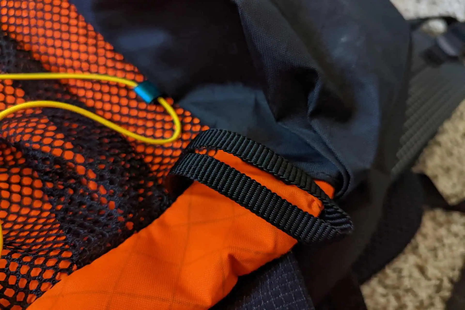 Close-up of an Atom Packs Pulse backpack showcasing textured black fabric with orange mesh, accented by a loop of black woven strap. A yellow cord with a blue plastic toggle adds a vibrant contrast, elevating the dynamic design of this premium hiking essential.