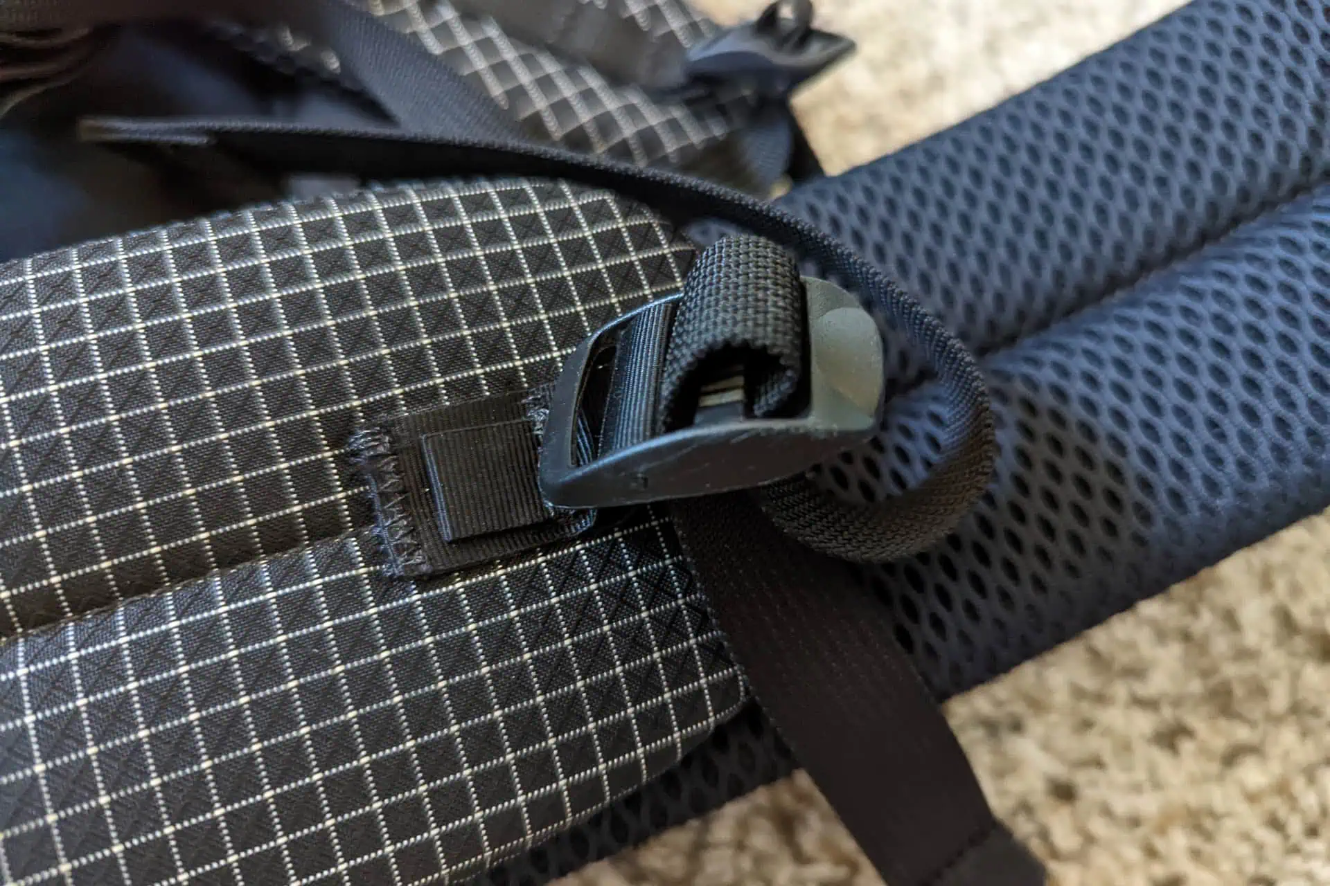 Close-up of the Atom Packs Pulse backpack strap, showcasing its checkered fabric. The strap features a sturdy plastic buckle and a section of black mesh padding. A textured carpet is visible in the background.