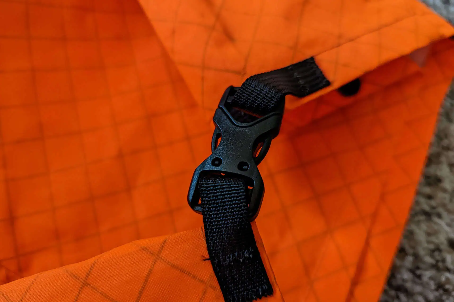 Close-up of a black plastic buckle fixed to a black strap on an orange fabric with a grid pattern, reminiscent of Atom Packs Pulse design. The fabric is slightly folded, and the buckle is partially closed.