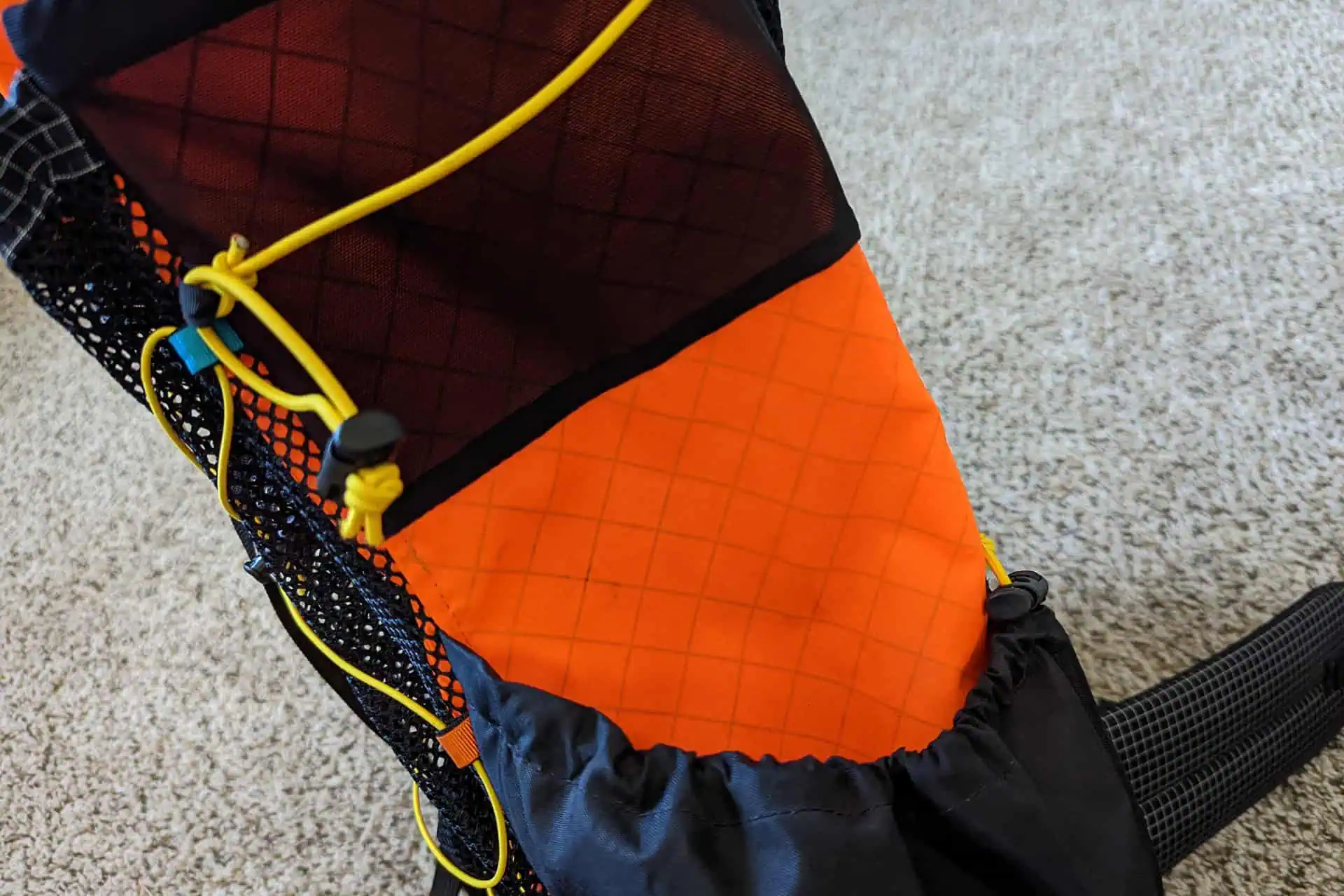 Close-up of an Atom Packs Pulse hiking backpack pocket with a grid pattern. It features yellow drawstrings, black mesh details, and a secure buckle resting on a gray surface.