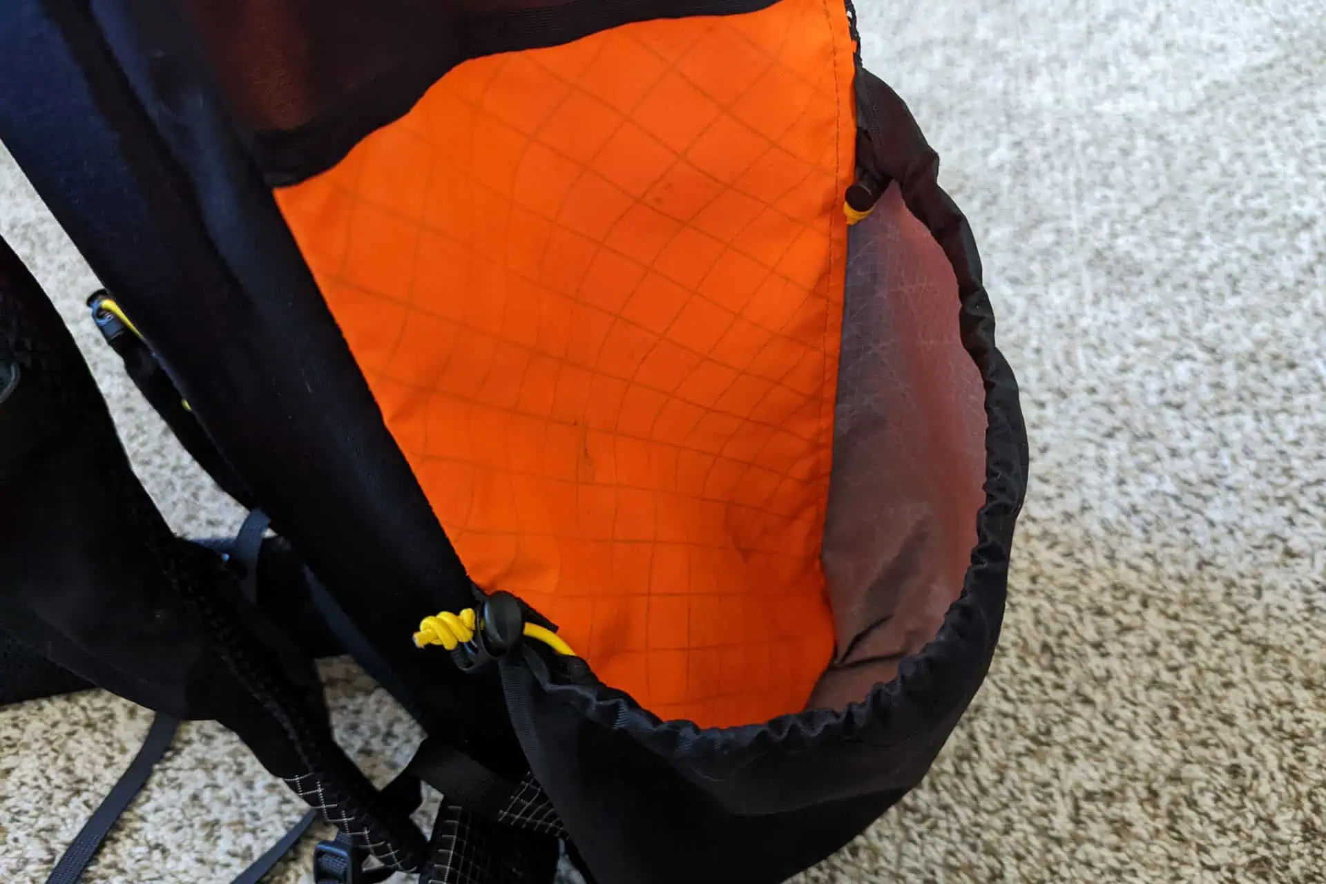 Close-up of an Atom Packs Pulse backpack in black and orange, featuring a side pocket. The orange fabric displays a grid pattern, with a yellow cord adjustment on the strap. Resting on a textured beige carpet, this bag combines practicality with sleek design.