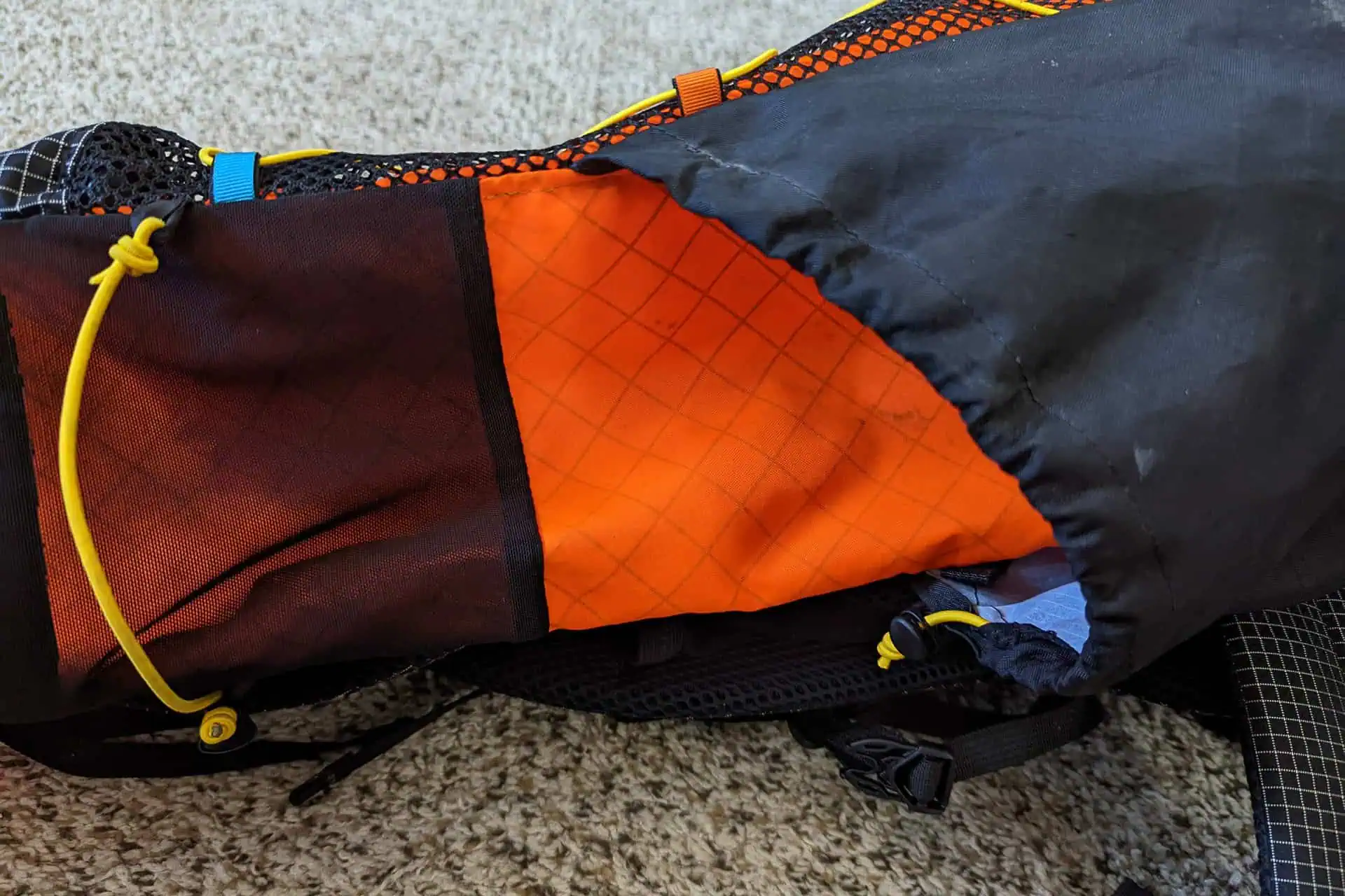 A backpack with an open pocket revealing an orange interior sits on a beige carpet. The exterior, reminiscent of the vibrant Atom Packs Pulse style, features black and red panels with yellow drawstrings. The pocket is lined with black mesh material for added flair.