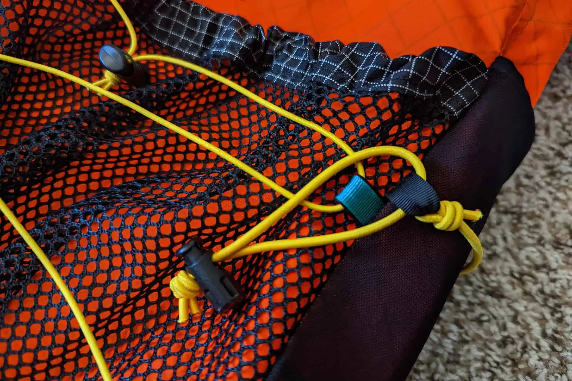 Close-up of an orange and black mesh Atom Packs Pulse backpack featuring yellow elastic cords threaded through loops and secured with toggles. Resting on a beige carpet, the backpack showcases its detailed stitching and stylish design elements.