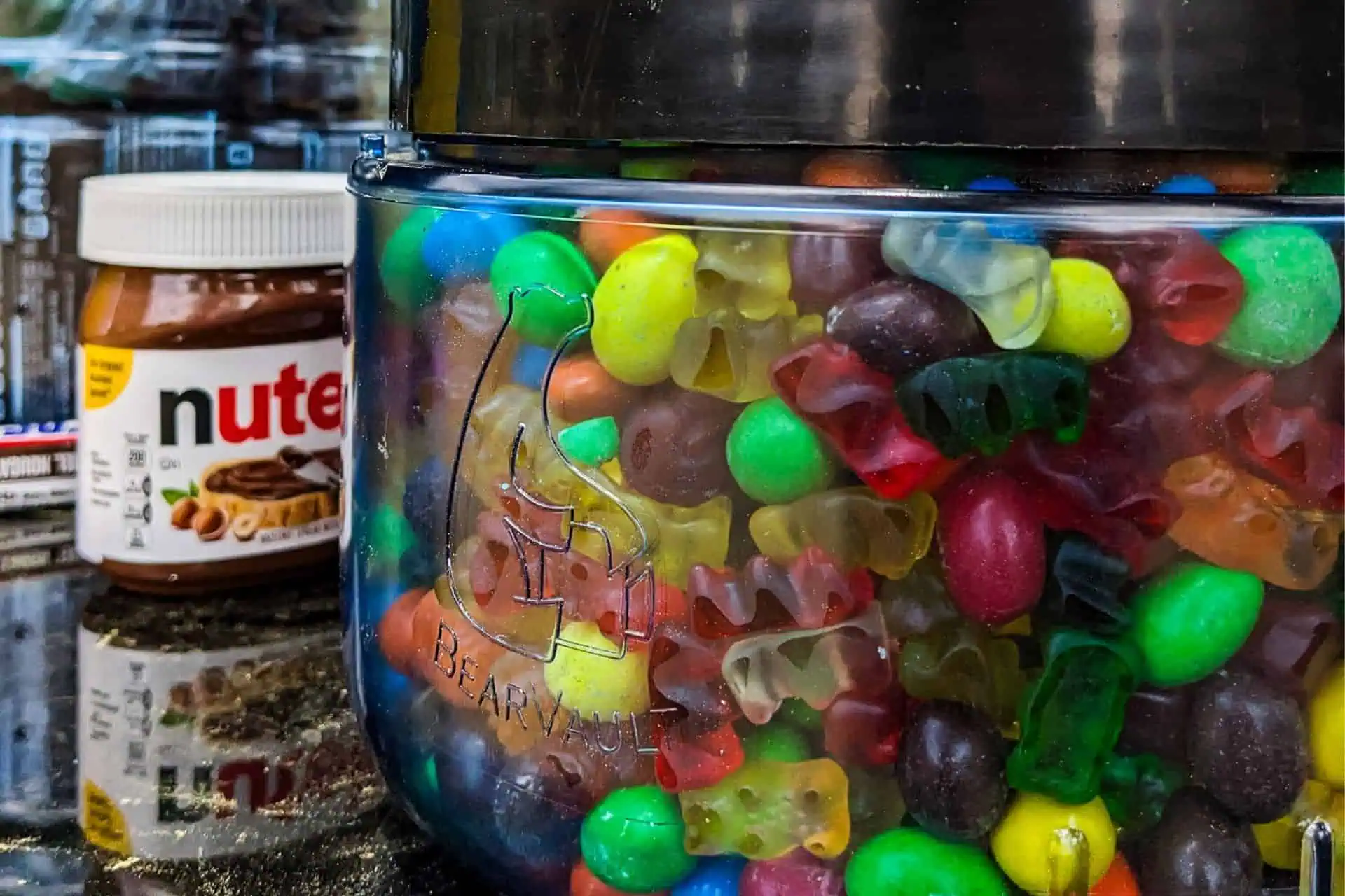 A close-up of a clear container filled with colorful gummy bears and candy, essential for any PCT resupply guide 2024. A jar of Nutella peeks from the background. The container features a bear logo and the word BearVault on it.