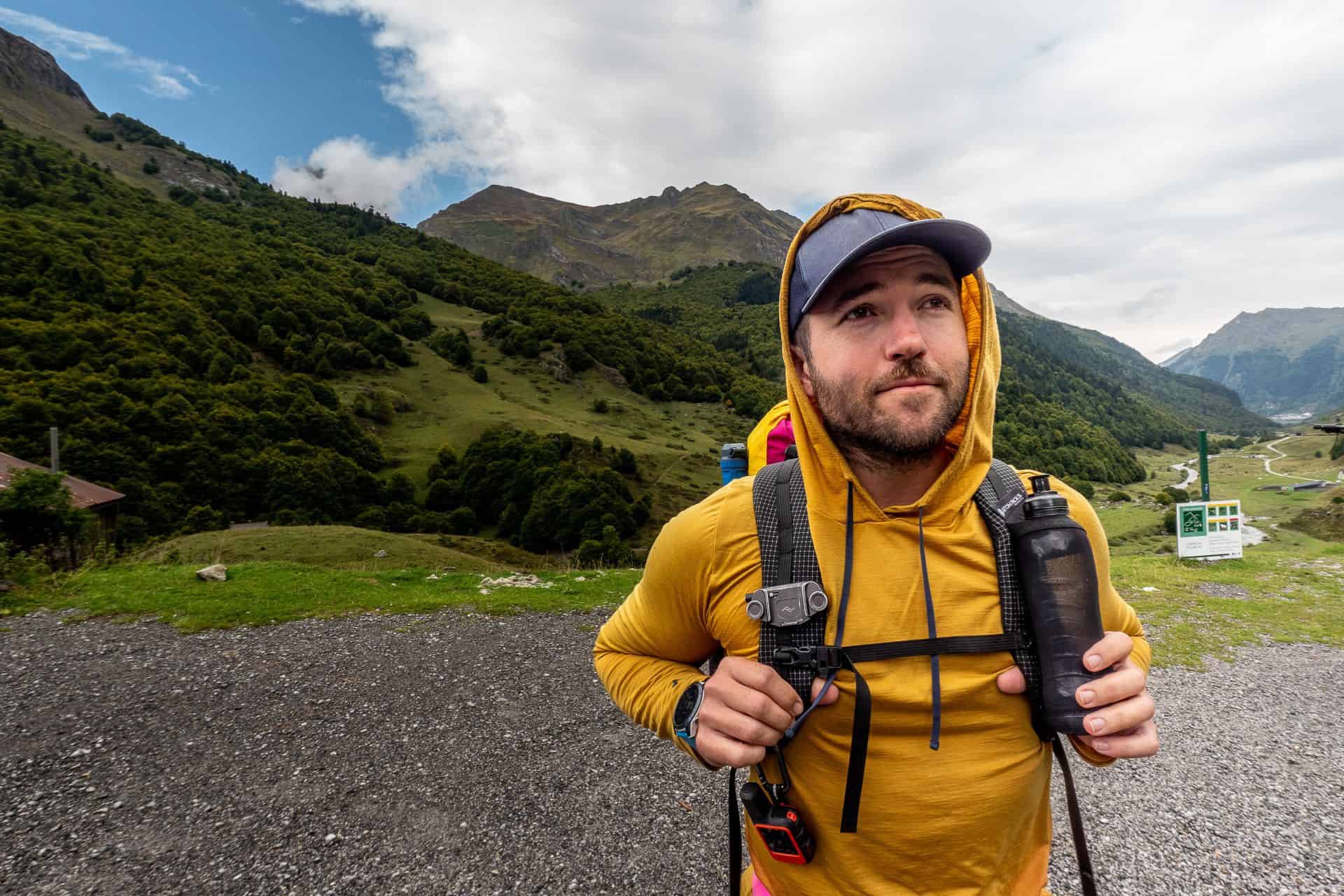 A man in a yellow hoodie and baseball cap stands outdoors, looking up thoughtfully, carrying his 2025 backpacking gear list. Behind him stretch green hills under a partly cloudy sky, promising adventure.