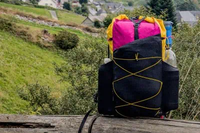 The Atom Packs Atom backpack, with a pink top and black body, features vibrant yellow crisscrossed cords and rests on a wooden ledge. Two water bottles are secured on the sides. In the background, a hilly landscape dotted with houses and trees stretches into the distance.