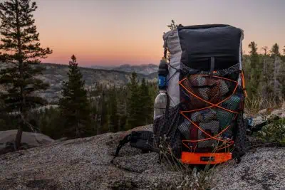 An Atom Packs Prospector backpack rests on a rocky surface with a scenic mountain view. The sky, softly lit by the setting sun, creates a warm glow around pine trees. The Prospector, filled with gear and featuring mesh pockets and orange accents, stands ready for adventure.
