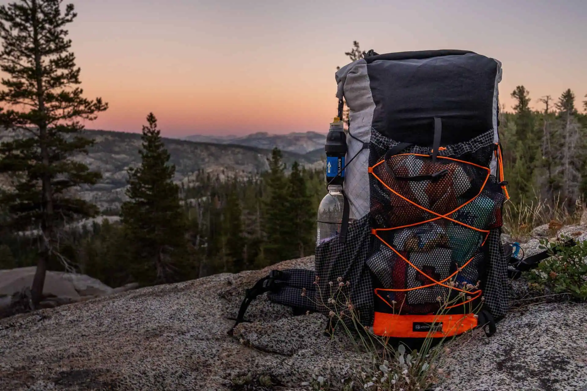 An Atom Packs Prospector backpack rests on a rocky surface with a scenic mountain view. The sky, softly lit by the setting sun, creates a warm glow around pine trees. The Prospector, filled with gear and featuring mesh pockets and orange accents, stands ready for adventure.