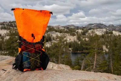 A vibrant Atom Packs Pulse backpack sits on a rocky surface overlooking a scenic landscape. In the background, rugged hills, pine trees, and a small lake are visible under a cloudy sky.