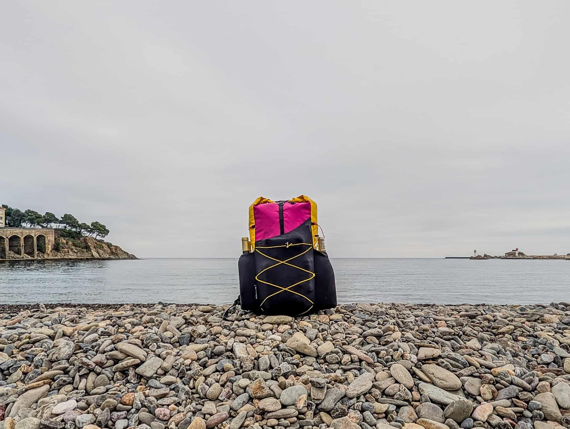 A backpack sitting on the shore of a bay beneath a cloudy sky.