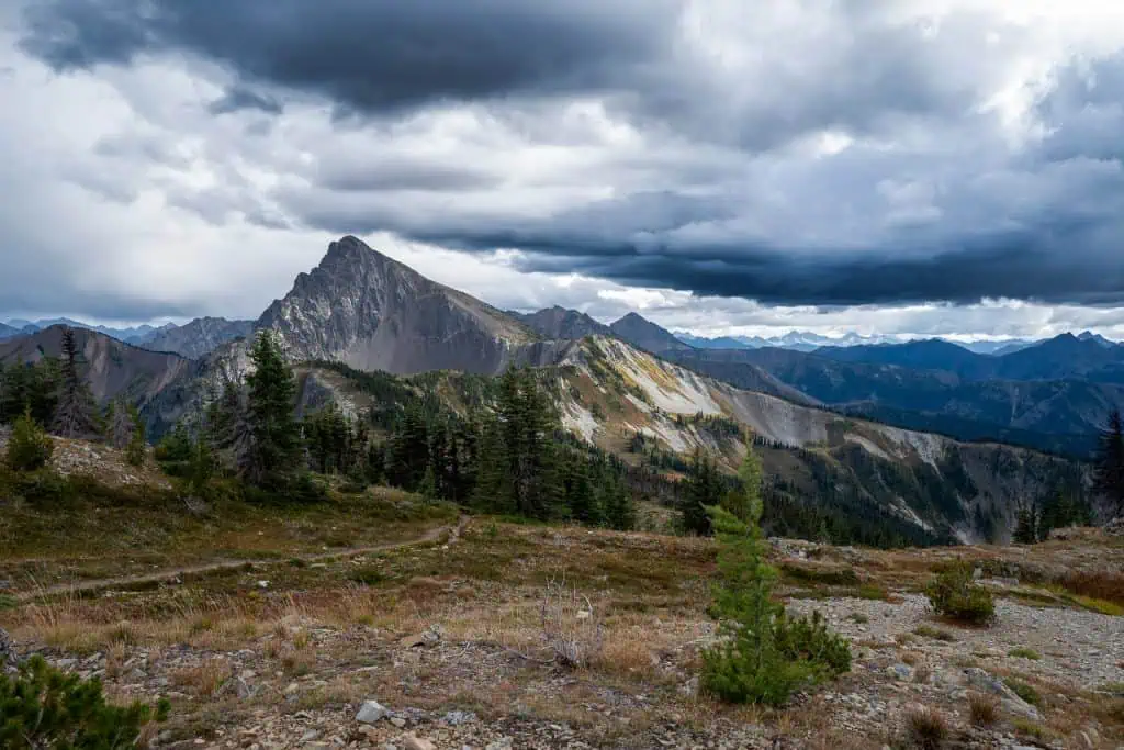 A dramatic mountain landscape under a cloudy sky unfolds like the setting of PCT horror stories 2024. The rugged trail winds through grassy terrain, leading to a prominent peak amid jagged mountains. Sparse trees and green patches add texture to this eerie yet breathtaking scene.