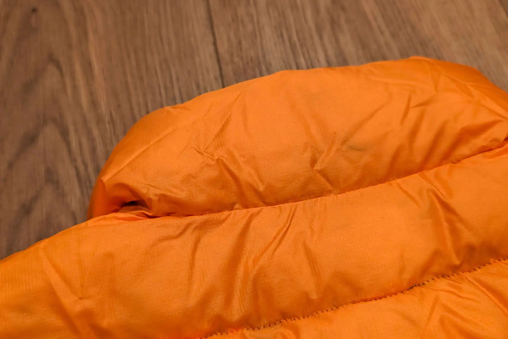 Close-up of an orange quilted fabric, possibly from a Feathered Friends Eos Vest, against a wooden background. The fabric has visible wrinkles and detailed stitching, capturing the essence of this cozy gear.