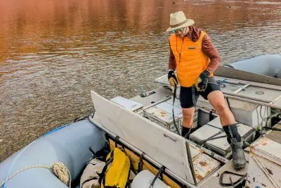 A person in a Feathered Friends Eos Vest, paired with shorts and a hat, stands on a raft near the riverbank, holding a rope. Sunglasses and boots complete their look. The raft brims with gear and supplies as calm waters reflect the majestic red rock cliffs in the backdrop.