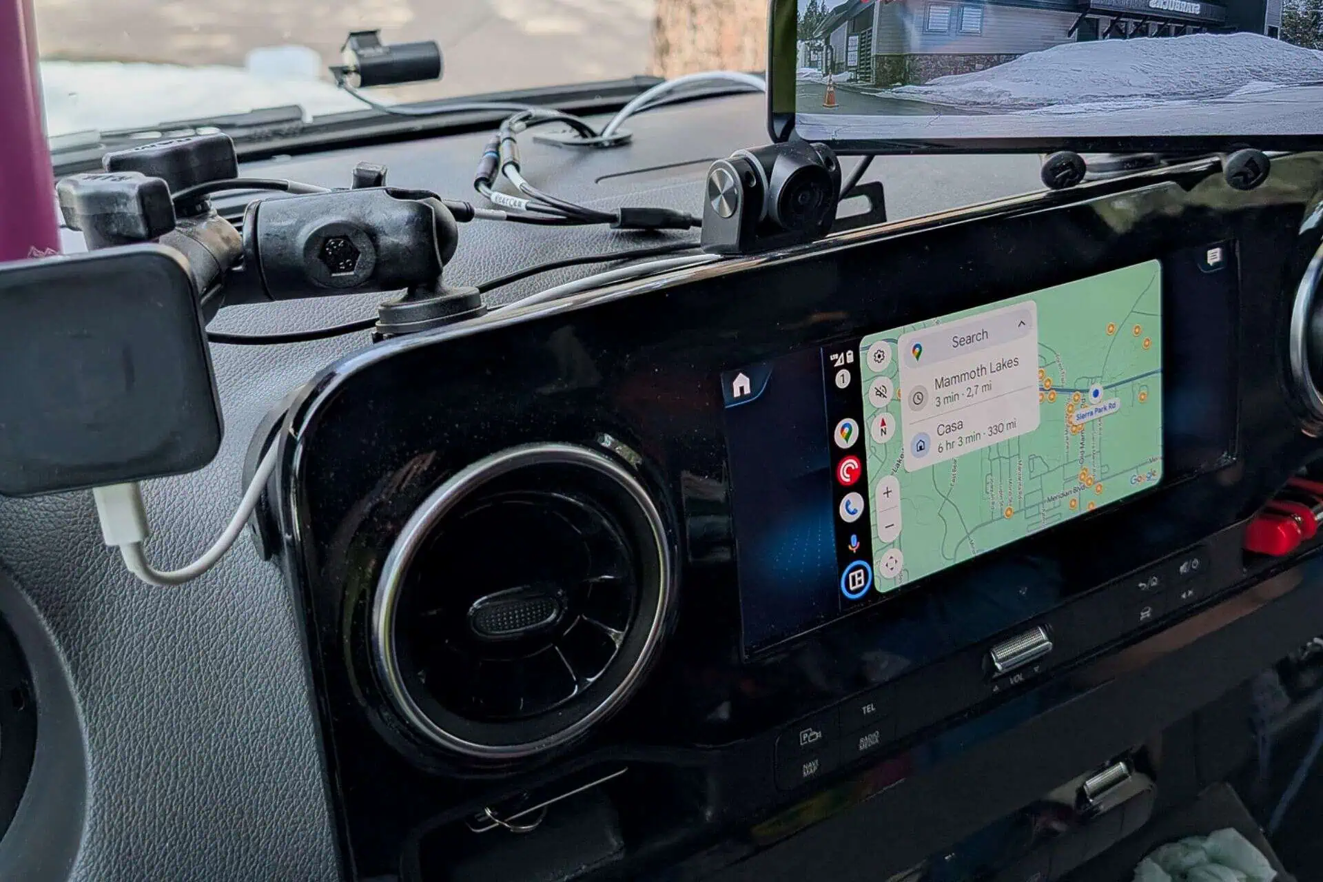 The car dashboard in the 2023 Mercedes Sprinter Van showcases a GPS map set for Mammoth Lakes on its digital screen, enhanced with Wireless Android Auto/Apple CarPlay. Several device holders and a phone charging cable adorn the setup, while a small rearview camera screen is perched at the top right.
