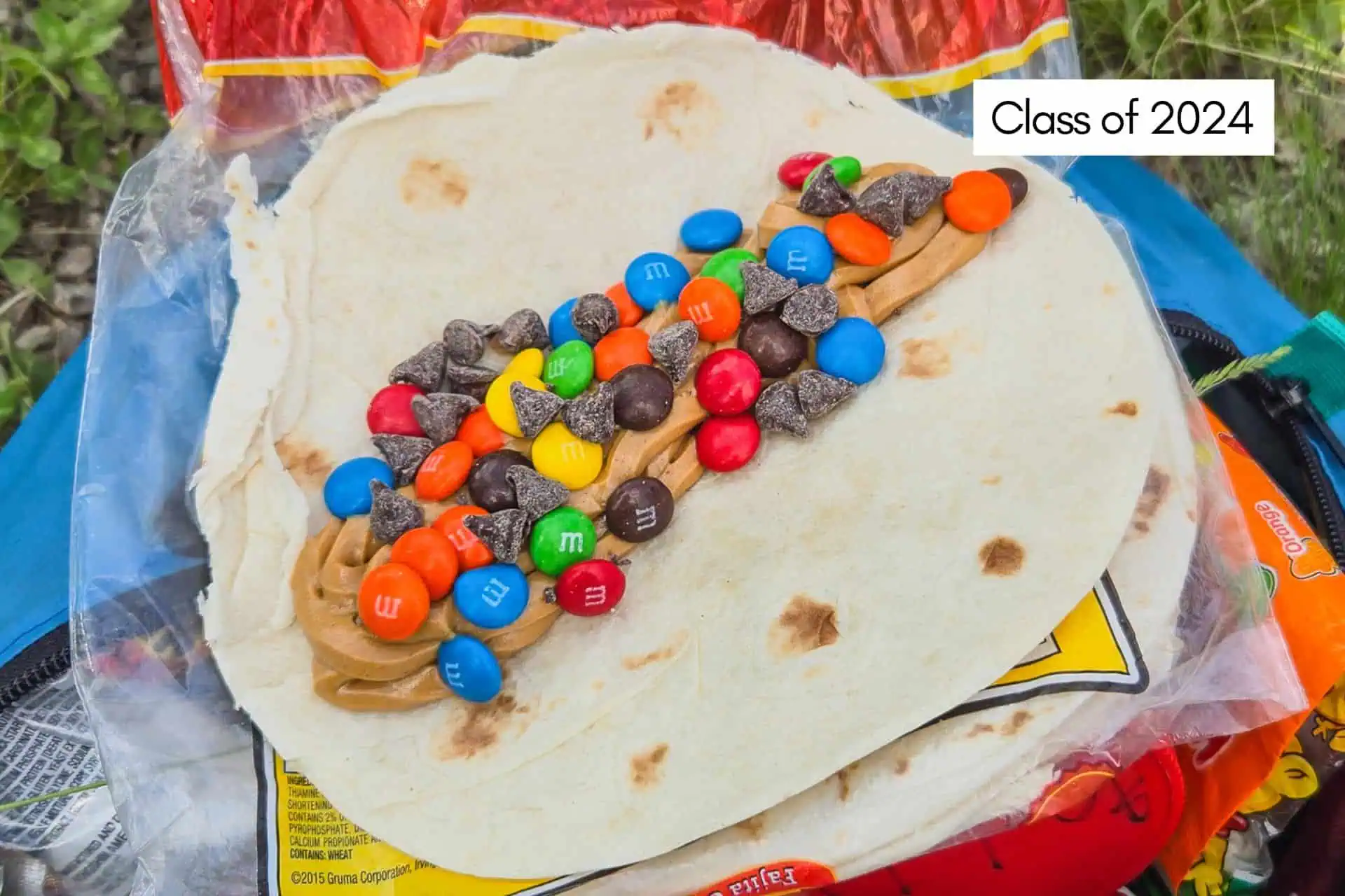 Tortilla with peanut butter, chocolate candies, and chocolate chips sits atop candy packaging against a backdrop of greenery. A label reads Class of 2024, hinting at a sweet send-off before diving into the upcoming adventures featured in the CDT Resupply Guide 2025.