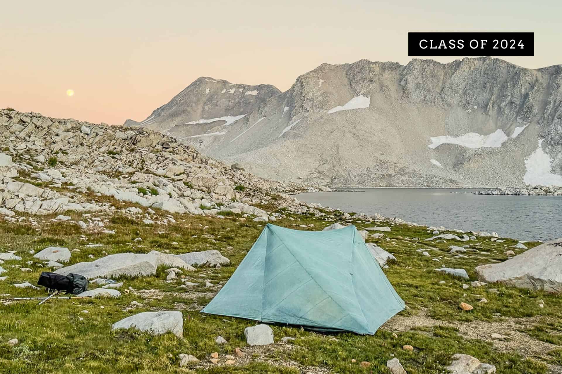 A blue tent from the JMT Gear Guide 2024 is pitched on a grassy patch surrounded by rocks, overlooking a serene mountain lake. Rugged mountains rise in the background under a pastel sunset sky. A sign proudly reads Class of 2024.