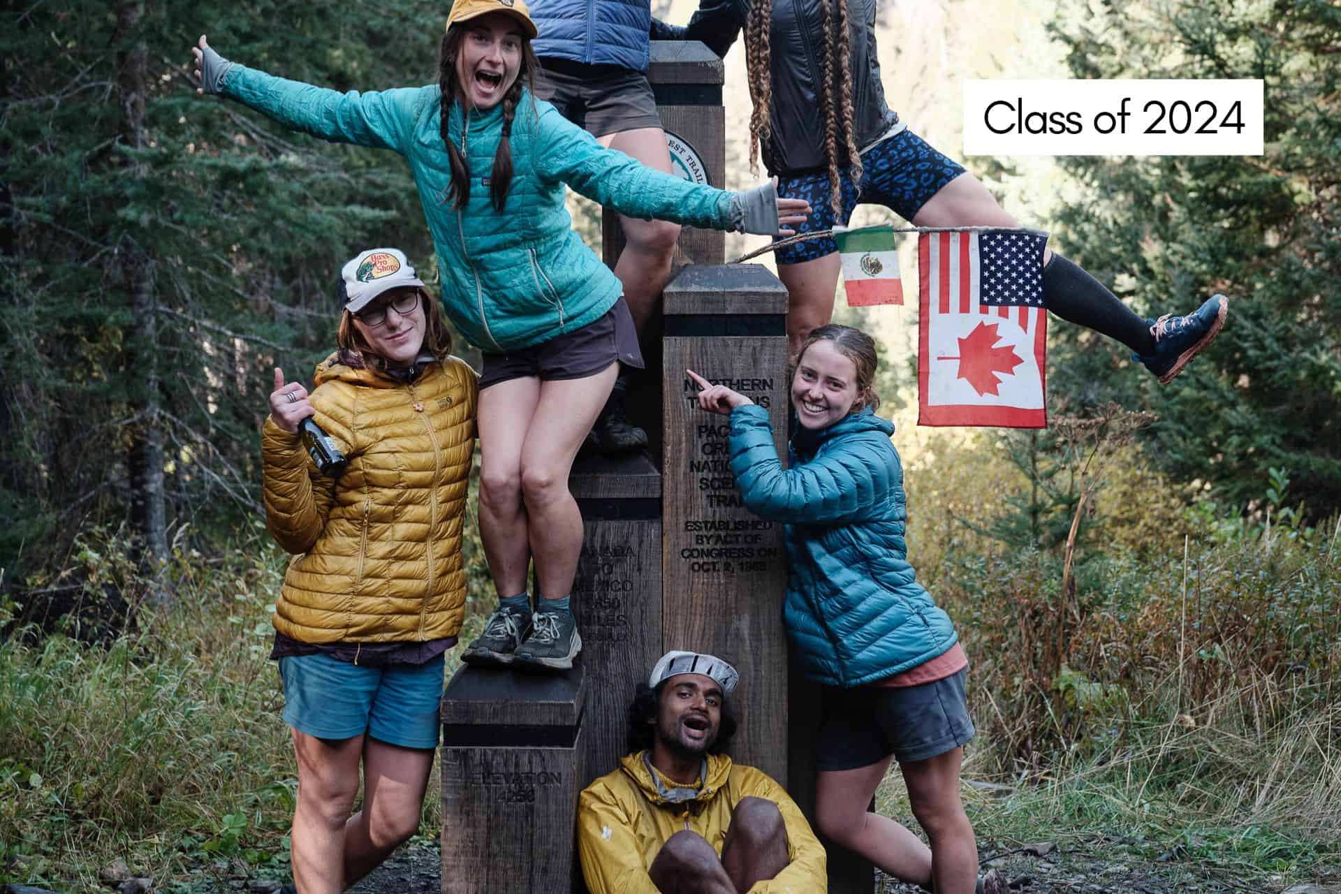 A lively group of six poses playfully on a monument adorned with flags from Mexico, the USA, and Canada. Dressed in outdoor gear and brimming with excitement for their 2024 Pacific Crest Trail hike, they stand beneath a Class of 2024 sign, surrounded by lush trees.