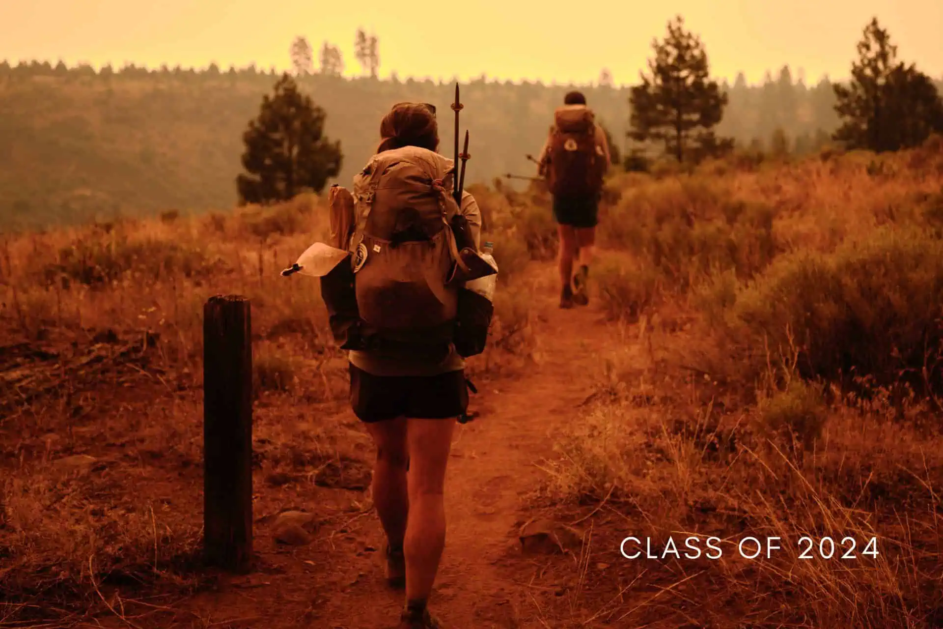 Two backpackers trek along a dirt trail through a dry, hilly landscape with trees, the sky hazy from wildfire smoke. As one hiker strides closer to the camera, Class of 2024 is emblazoned in the corner, hinting at untold PCT horror stories lurking ahead.