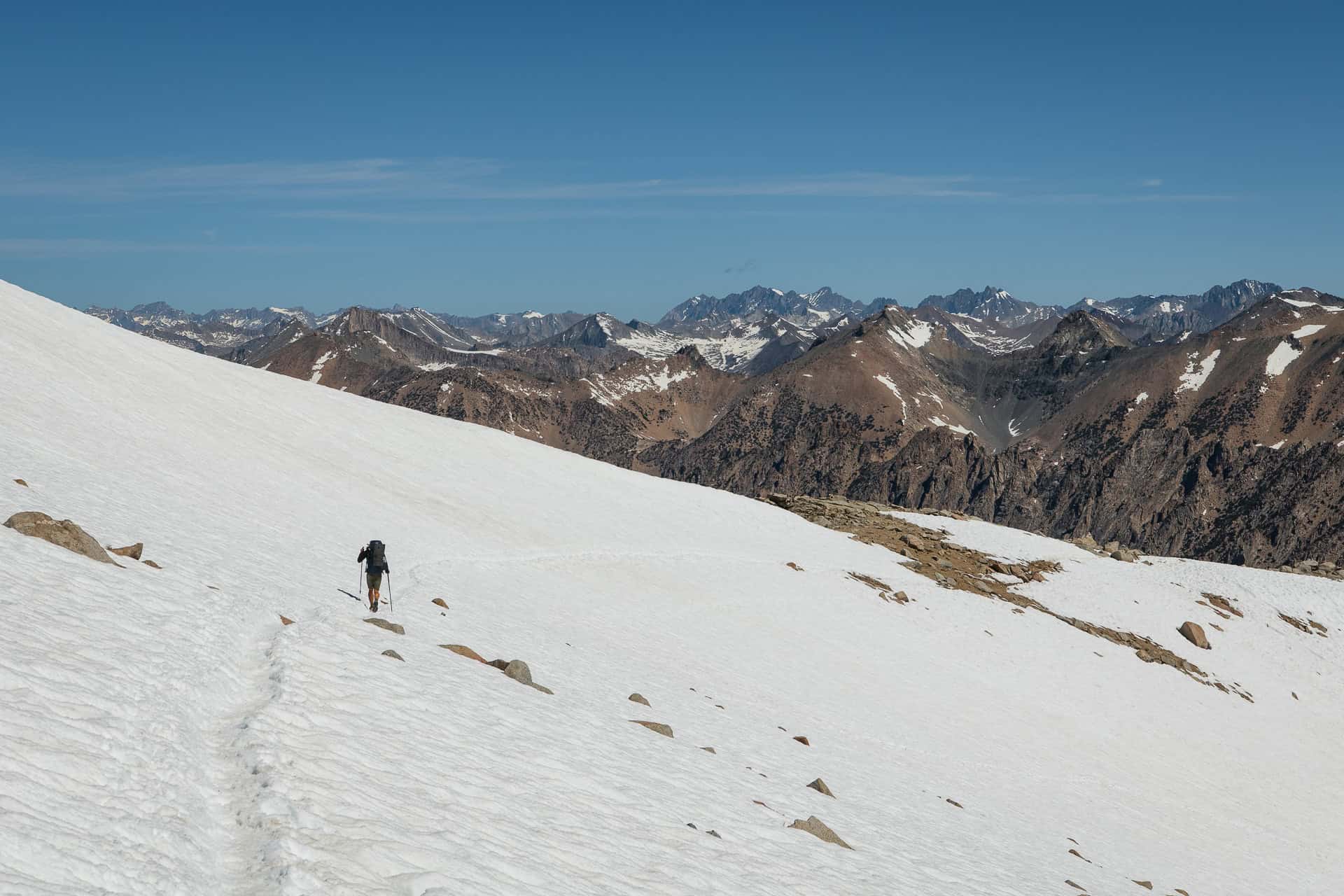 A lone hiker with a backpack traverses a vast, snowy mountain slope under a clear blue sky. Rugged, rocky peaks reminiscent of the Pacific Crest Trail are visible in the background, creating a dramatic alpine landscape perfect for adventure in 2025.