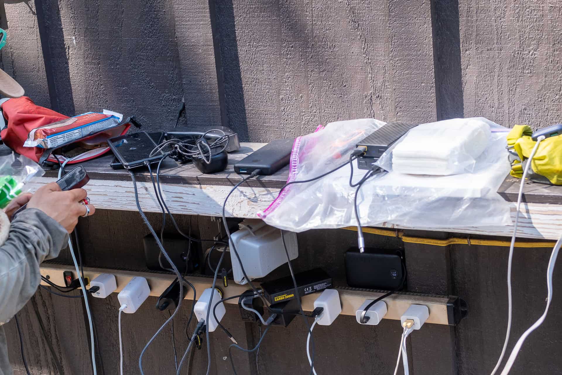 Amidst the chaos of managing multiple electronic devices and charging cables on a wooden shelf outdoors, a person juggles through what feels like one of their worst moments. Various chargers are plugged into a power strip, sharing space with gloves and a plastic bag amidst the hubbub.