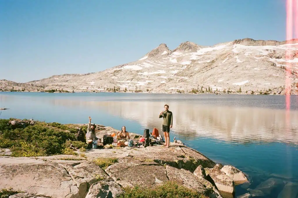 Four people relax on rocky terrain by a lake in a mountainous area—one of 2024s best PCT moments. Three wave from the ground while one stands drinking. Snow-capped peaks and clear water reflect the bright, sunny sky, with camping gear scattered nearby.