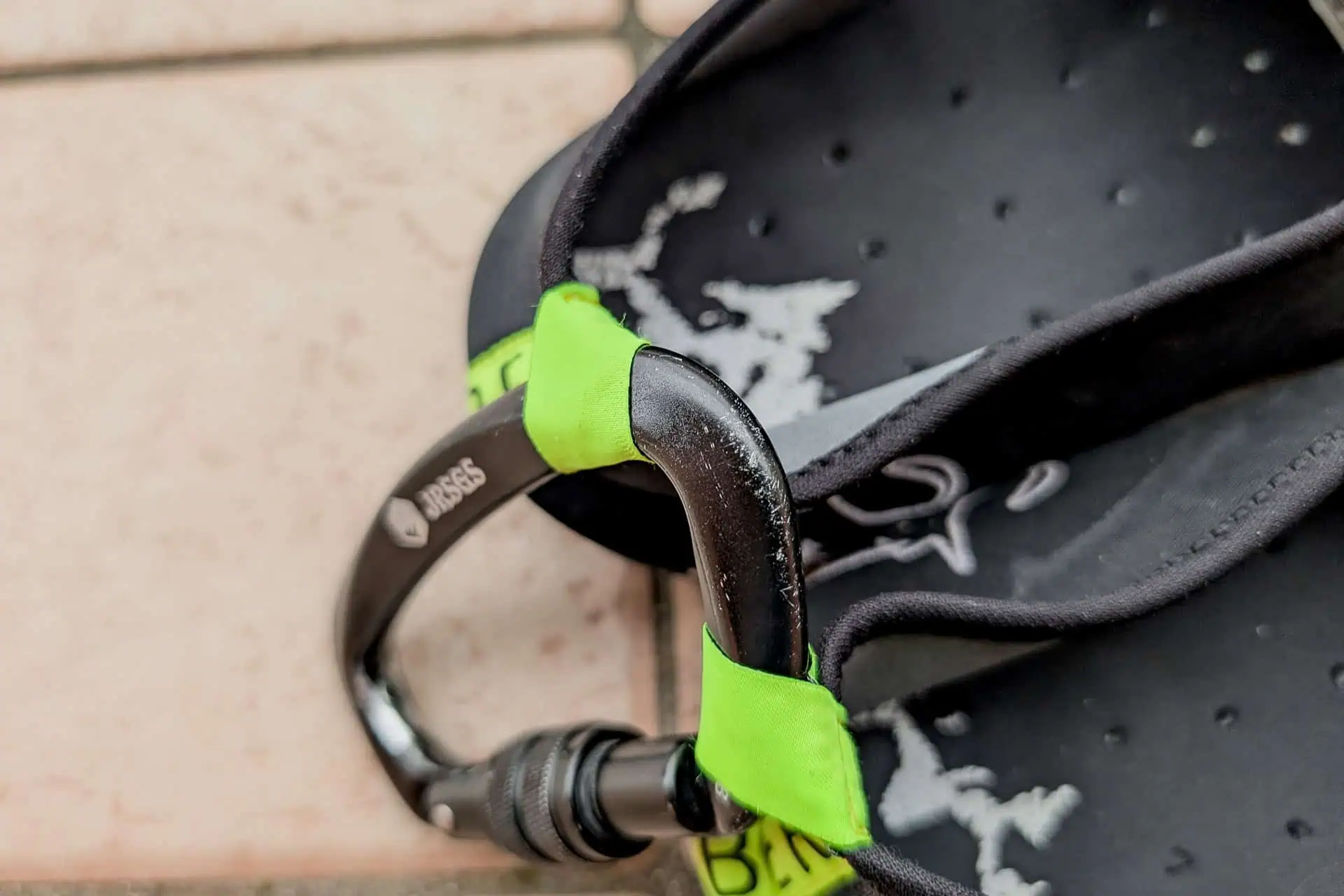 Black sandals with green accents, branded as BERT Shoes, are attached together by a black carabiner on a tiled floor.