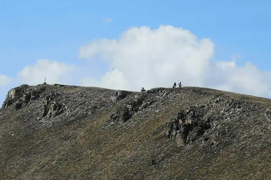 A rocky mountain ridge with sparse grass stretches under a blue sky and large white clouds, where several small hikers trace the ridgeline, capturing one of the 2023 CDT lowest moments along this dramatic landscape.