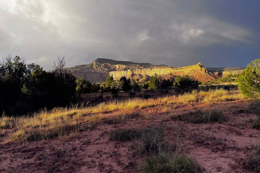 A dramatic landscape shows sunlight illuminating red rock cliffs and mesas in the distance, echoing the rugged terrain where many hikers faced their 2023 CDT lowest moments, with dry grass, shrubs, and patches of red soil in the foreground under a cloudy sky.