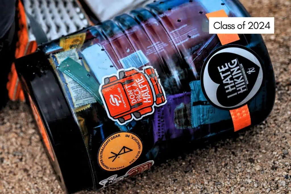 A close-up of a transparent container filled with various colorful items. It features several stickers, including I HATE HIKING, a circular symbol, and text reading Class of 2024. Perfect for your JMT resupply essentials, the background reveals a textured surface.