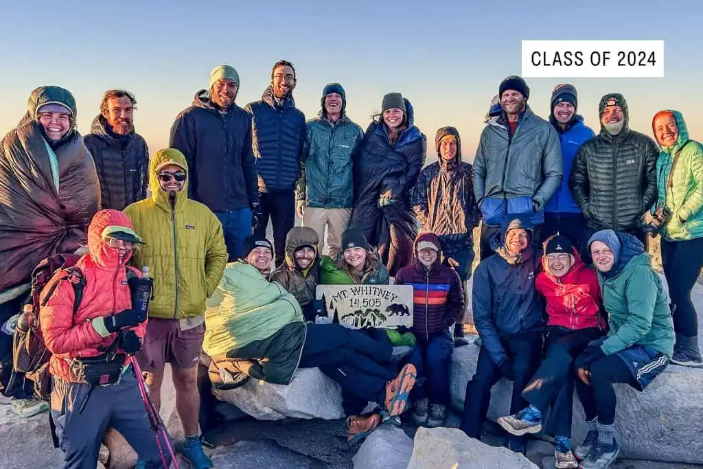 A group of hikers in outdoor gear smile and pose together on rocky terrain, holding a sign that reads Mt Whitney 14,505. A label in the corner reads Class of 2024—one of the 2024 best pct moments under a clear sky.