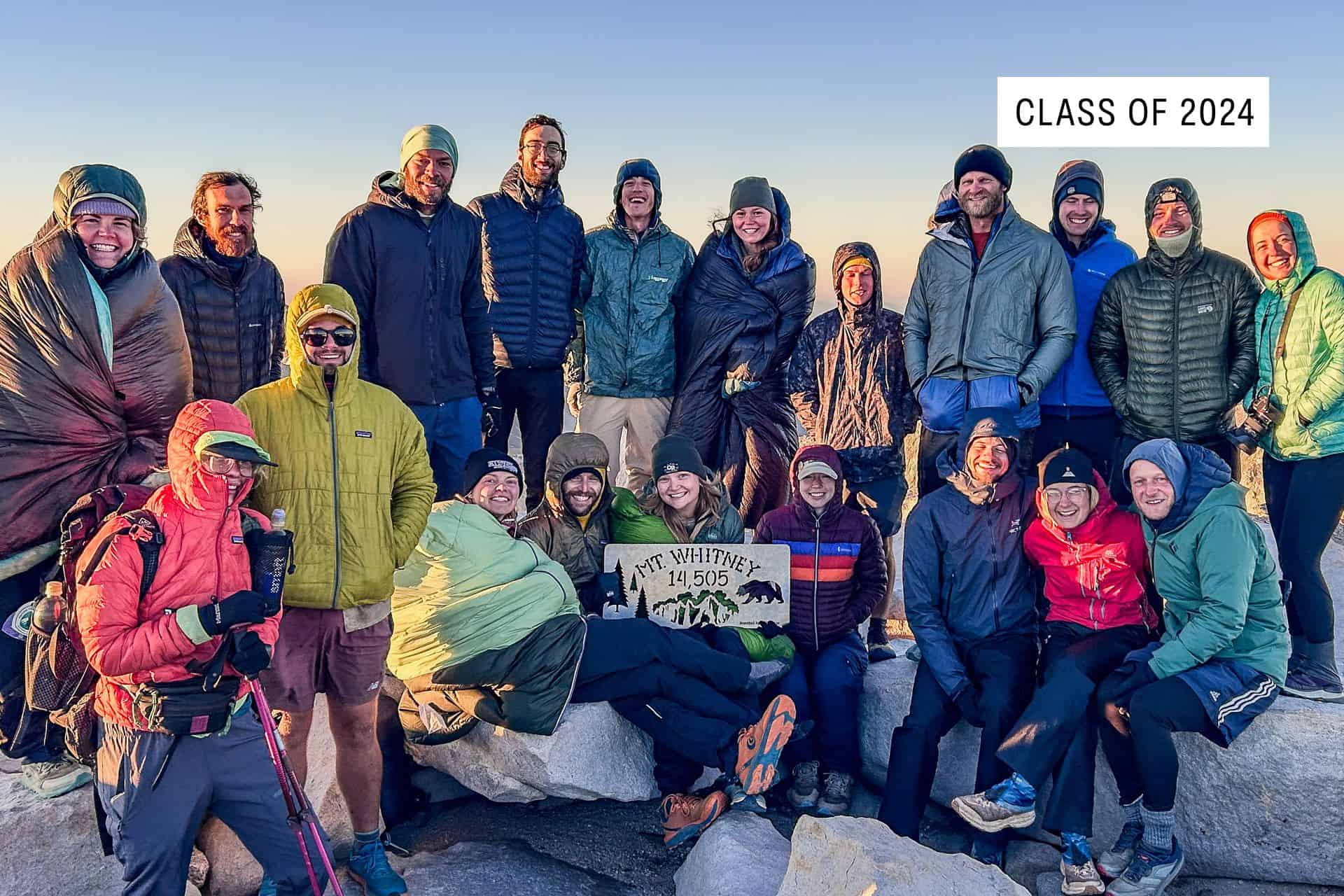 A group of hikers in outdoor gear smile and pose together on rocky terrain, holding a sign that reads Mt Whitney 14,505. A label in the corner reads Class of 2024—one of the 2024 best pct moments under a clear sky.