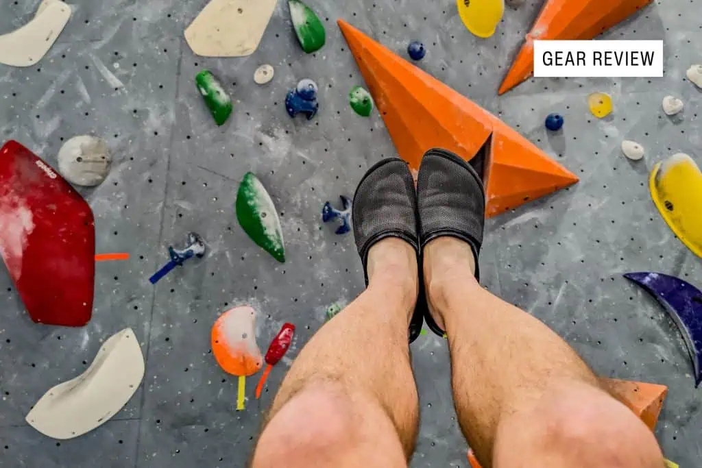 A close-up of a climbers lower legs wearing black BERT Shoes on a colorful indoor rock climbing wall. The wall features various holds in shapes like triangles and circles. A Gear Review label is in the upper right corner.