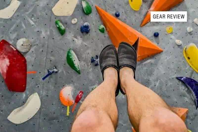 A close-up of a climbers lower legs wearing black BERT Shoes on a colorful indoor rock climbing wall. The wall features various holds in shapes like triangles and circles. A Gear Review label is in the upper right corner.