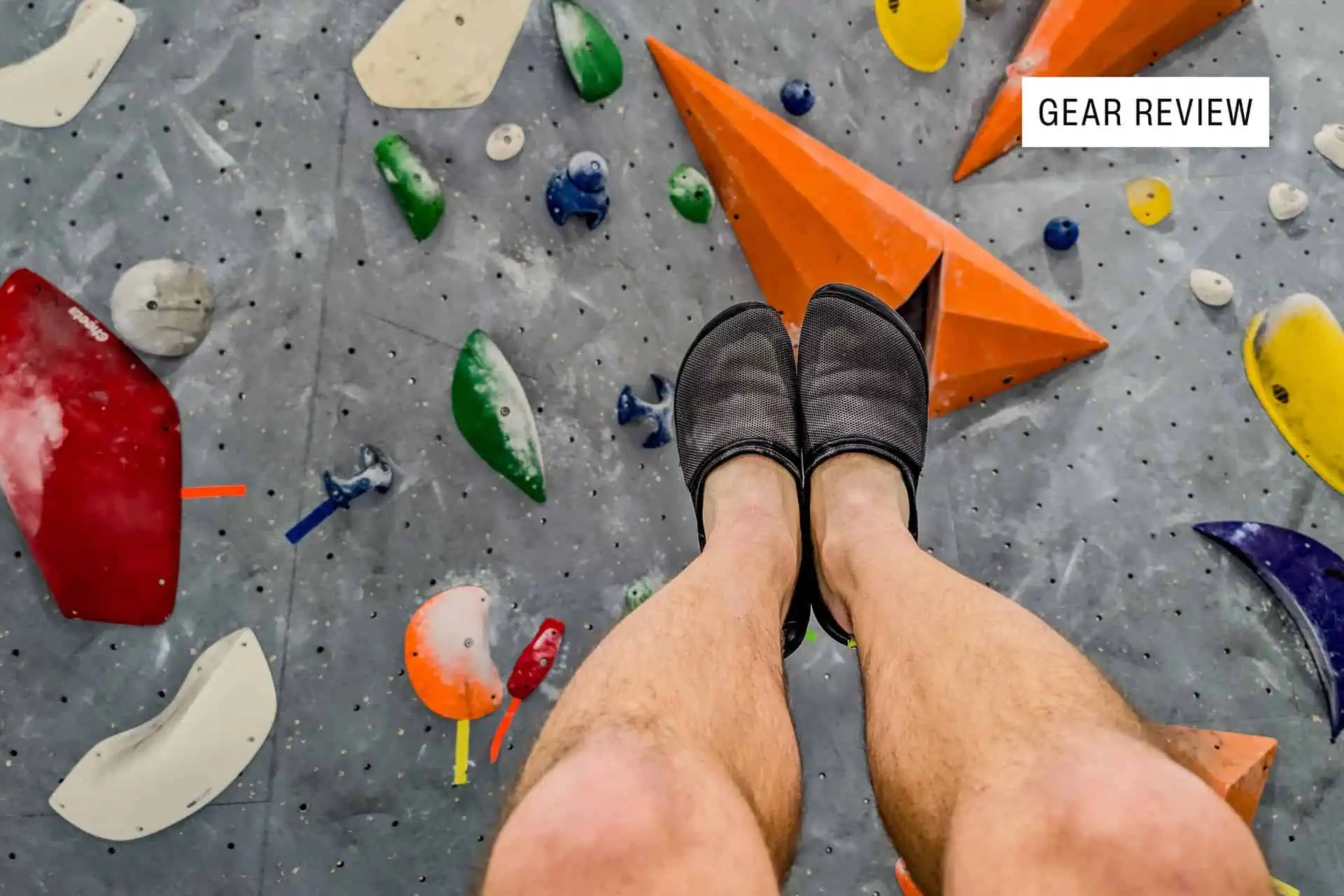 A close-up of a climbers lower legs wearing black BERT Shoes on a colorful indoor rock climbing wall. The wall features various holds in shapes like triangles and circles. A Gear Review label is in the upper right corner.