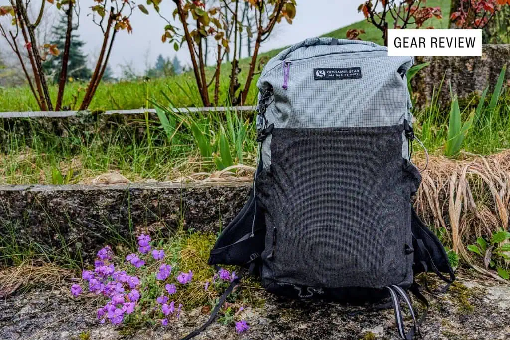 A gray and black Gossamer Gear Grit 28 backpack rests on a stone surface, accented by a patch of purple flowers. In the backdrop, green grass and shrubs complete the scene. A Gear Review label is at the top right, capturing attention.