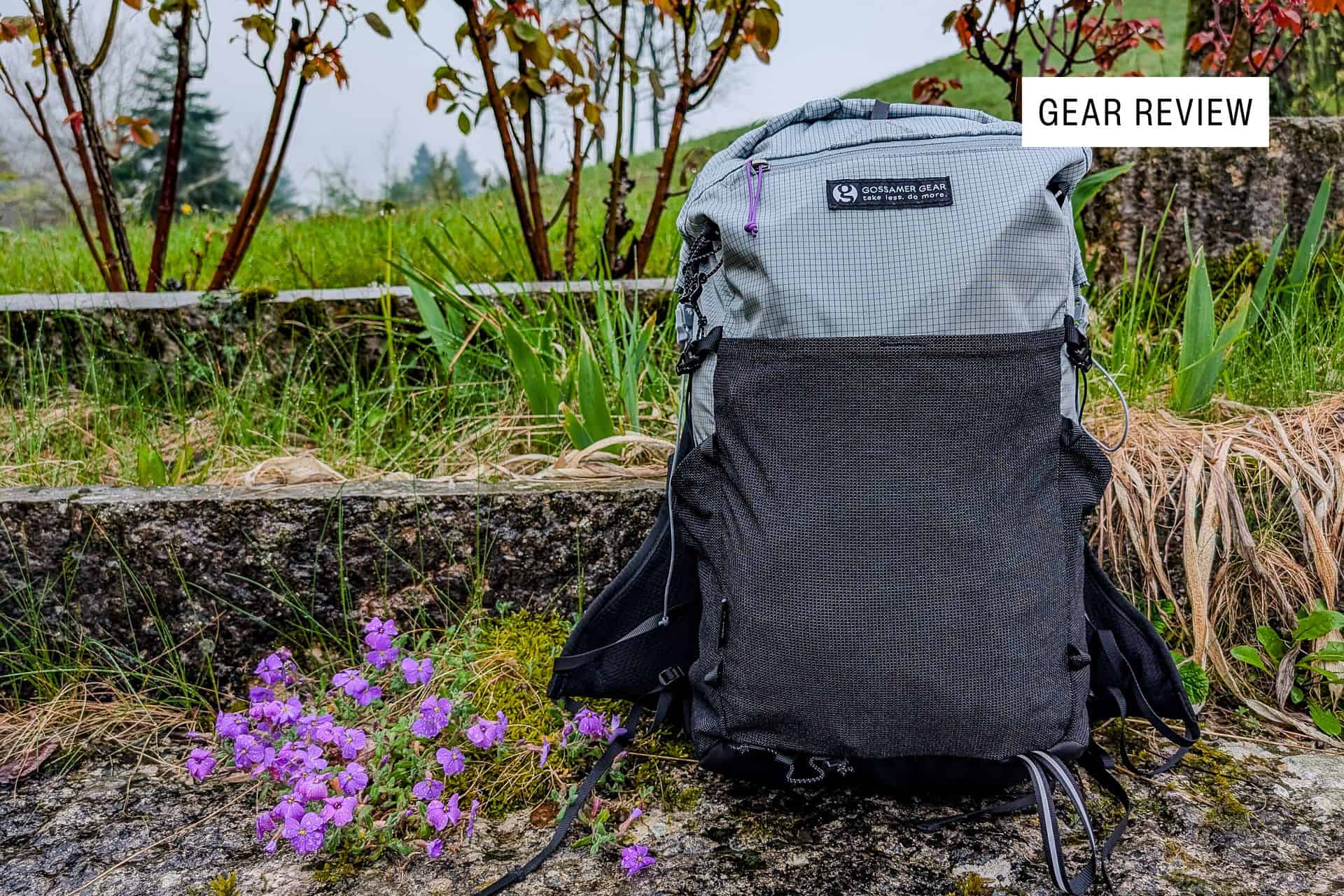 A gray and black Gossamer Gear Grit 28 backpack rests on a stone surface, accented by a patch of purple flowers. In the backdrop, green grass and shrubs complete the scene. A Gear Review label is at the top right, capturing attention.