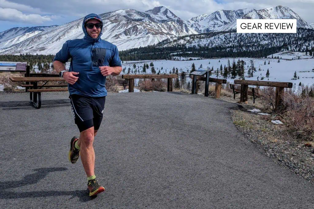 A person in Ridge Merino Solstice Pro athletic gear is jogging on a paved path with snowy mountains and pine trees in the background. A table and benches are on the left. The sky is partly cloudy. Text in the top right reads GEAR REVIEW.